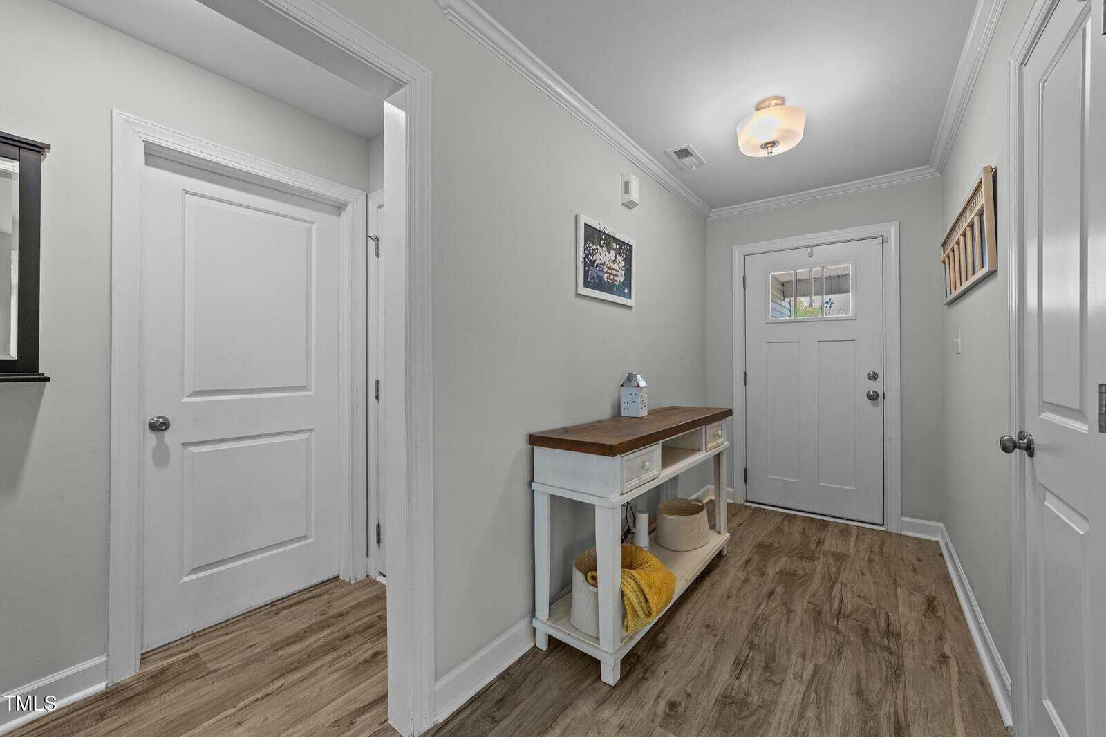 A hallway with light gray walls, wood flooring, and a white front door. There is a white console table with baskets and decor, and framed art on the walls. Two closed doors are visible on the left and right.