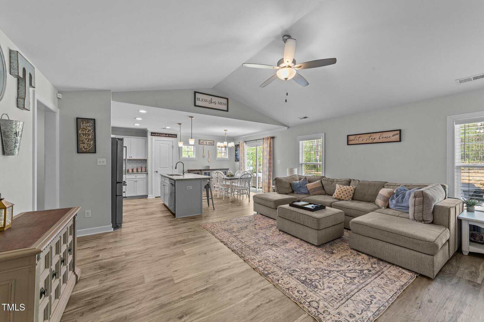 Open living room and kitchen with light wood floors, gray sectional sofa on a patterned rug, ceiling fan, kitchen island, dining table, and large windows with blinds letting in natural light.