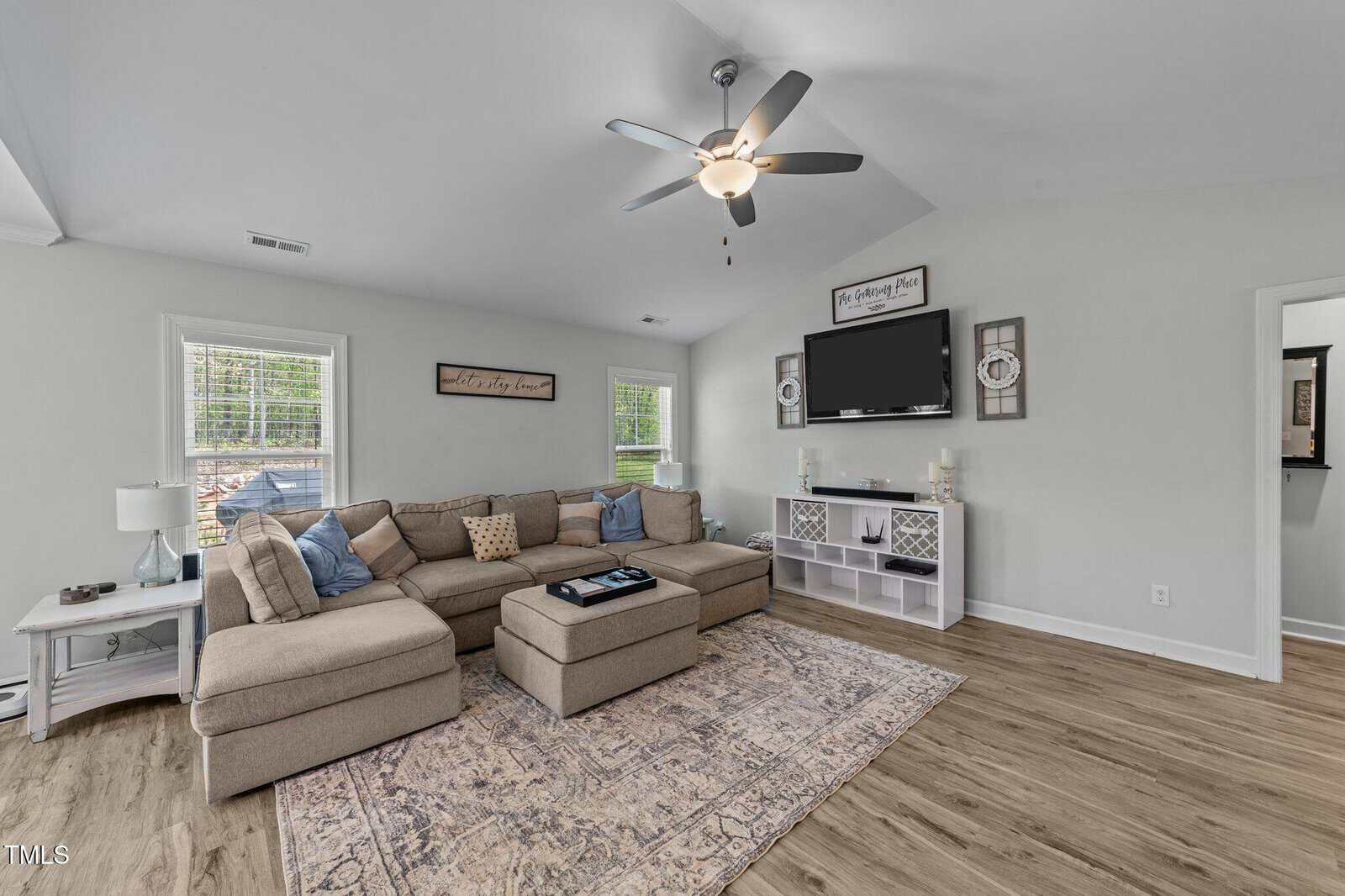 A living room with a sectional sofa, ottoman, and area rug on wood flooring. A ceiling fan and TV are mounted on light gray walls, with shelves and decor. Two windows let in natural light.