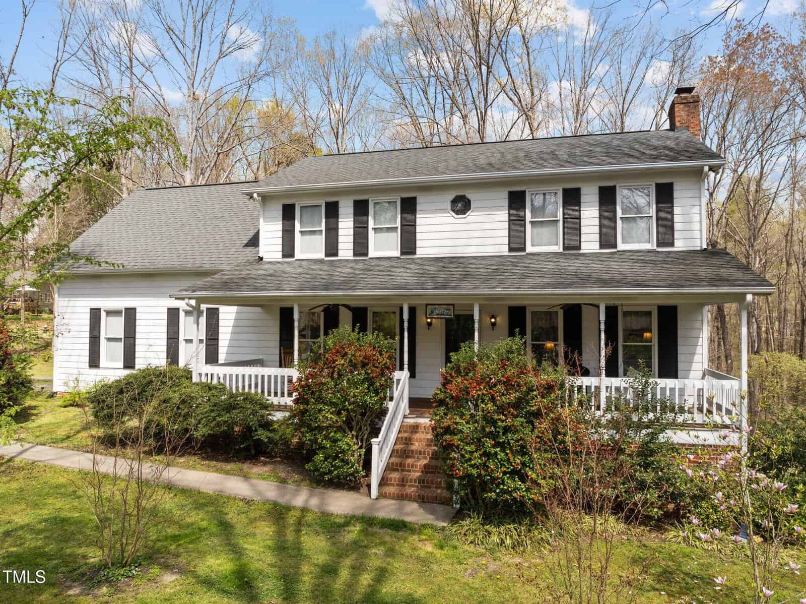 Located at 323 E Stratford Drive in Henderson, this charming two-story white house features dark shutters and a welcoming front porch surrounded by lush bushes and trees. A gray roof tops the structure, complemented by a brick chimney. The well-maintained lawn boasts a pathway leading to the entrance.