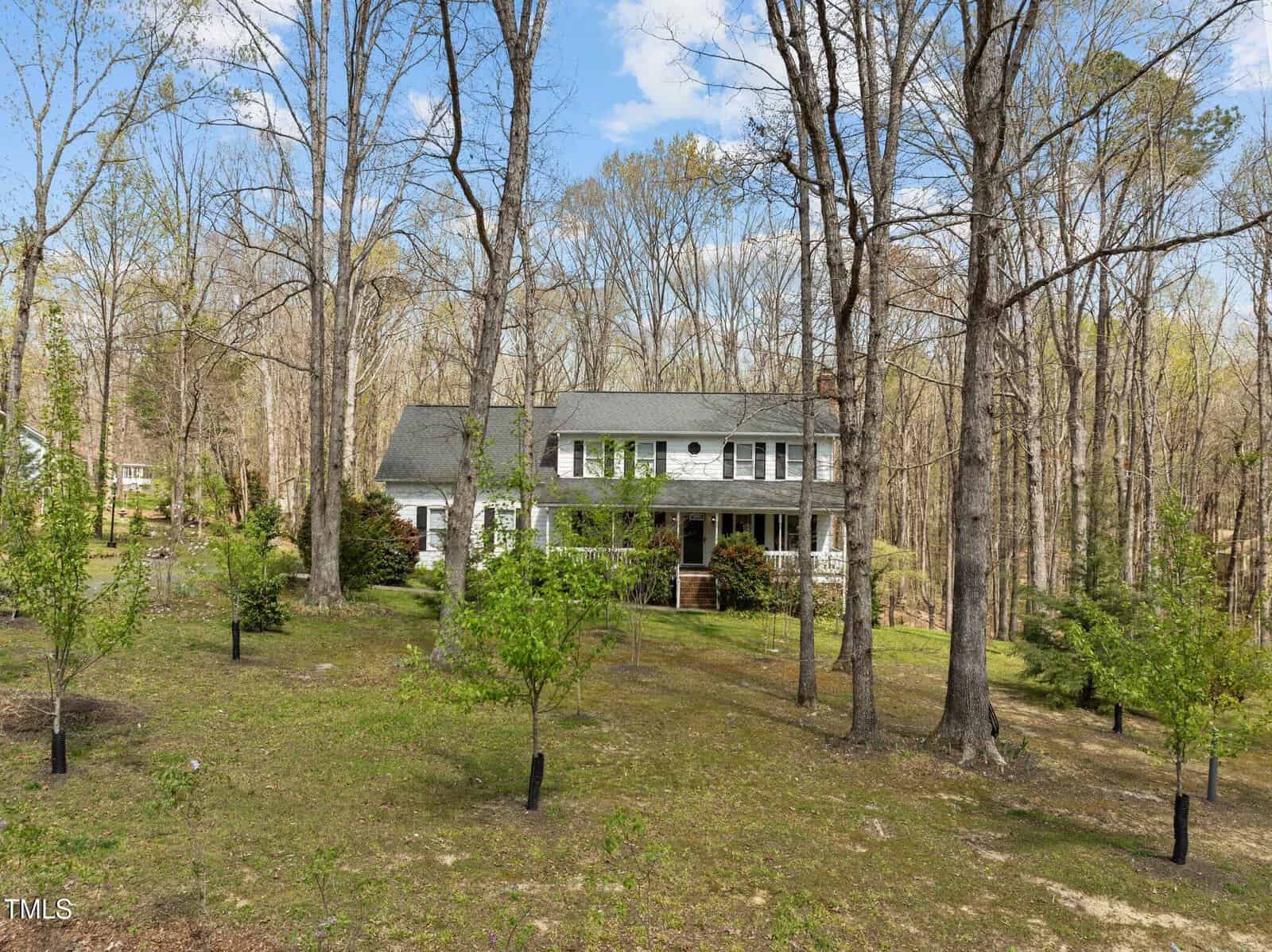Two-story white house with a gray roof, set in a wooded area surrounded by trees and a green lawn. The house features a porch and several small trees planted in the front yard under a clear blue sky.