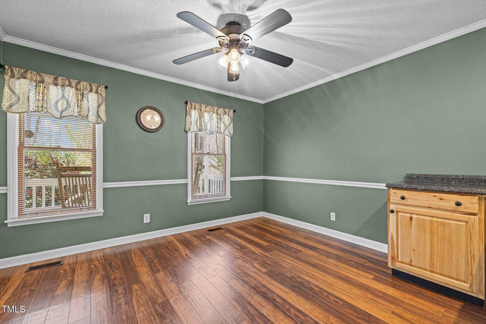 An empty room with green walls and wood flooring. Two windows have patterned curtains. A lighted ceiling fan is centrally positioned above. A wooden cabinet with a dark countertop is on the right wall. A small wall clock is between the windows.