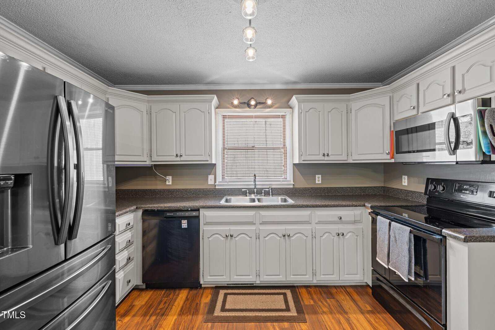 A kitchen with white cabinetry, dark countertops, and stainless steel appliances, including a refrigerator, dishwasher, oven, and microwave. A window above the sink lets in natural light. The wood floor features a small rug.