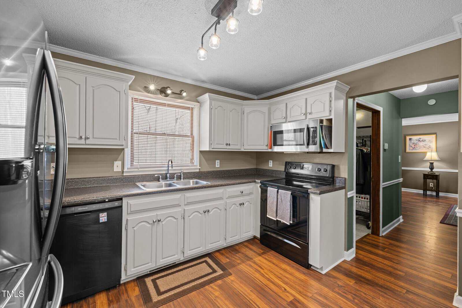 A kitchen with white cabinets, dark countertops, and wood flooring. It features a black stove, a modern ceiling light fixture, a double sink under a window with blinds, and a stainless steel refrigerator. A doorway leads to another room.
