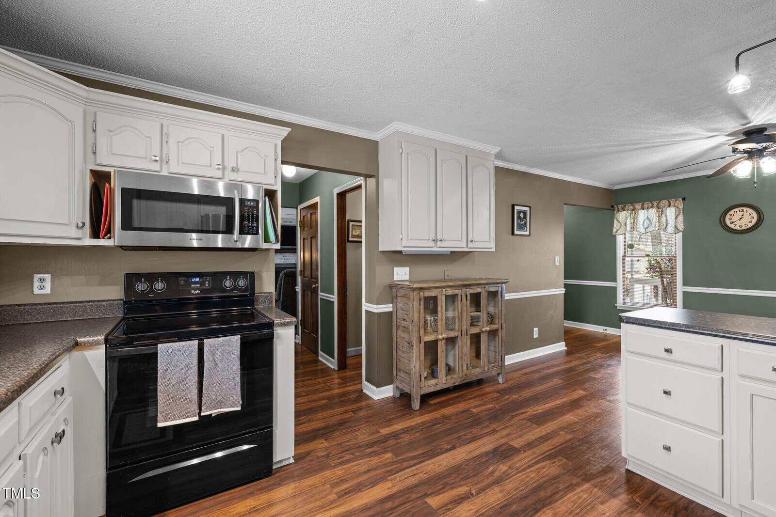 A kitchen with white cabinets and a green accent wall. It has stainless steel appliances, including a microwave and a stove. Wooden floors and a ceiling fan are visible. There is a wooden cabinet and a window with checked curtains in the dining area.
