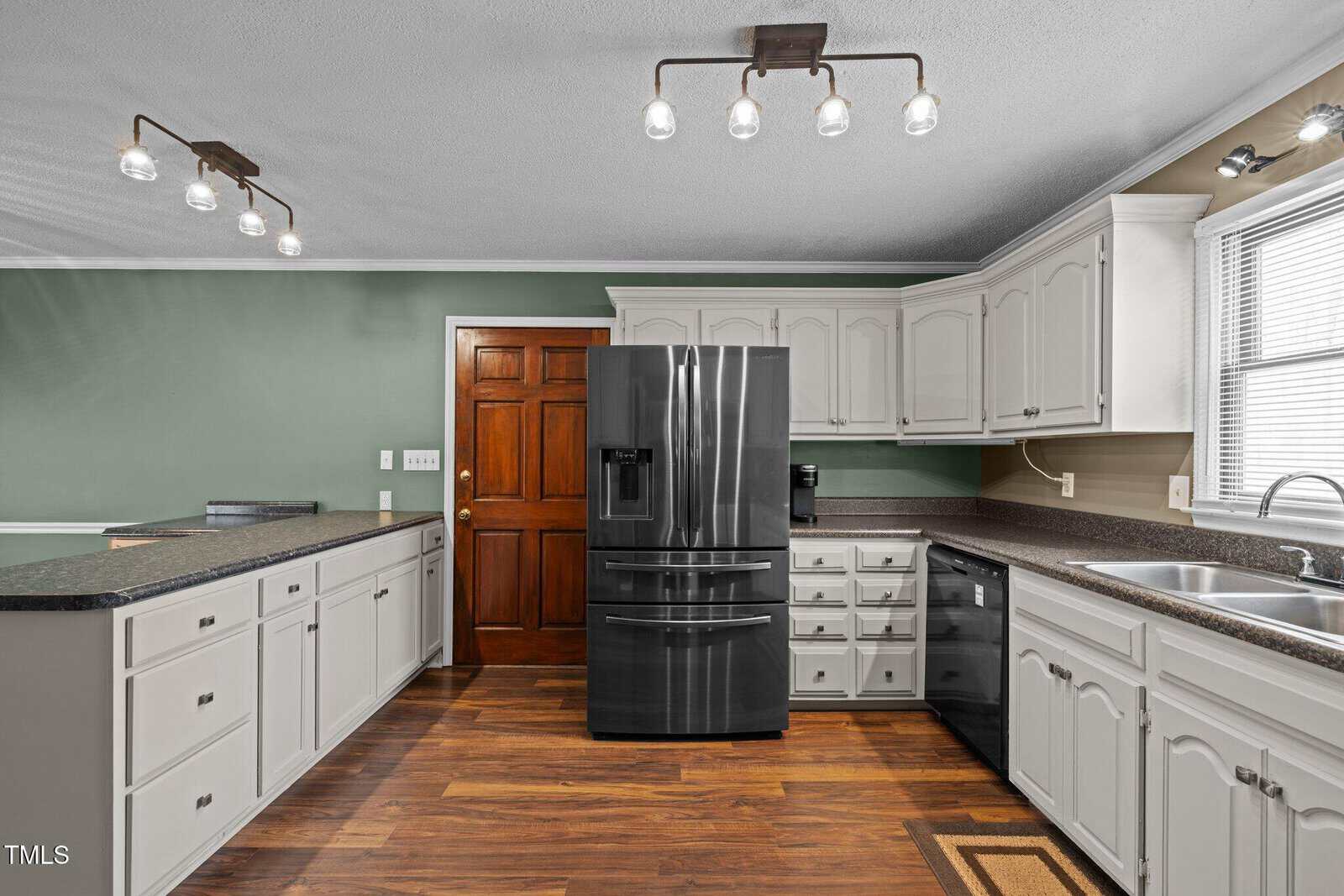 A kitchen with green walls, white cabinets, and wood flooring. Stainless steel refrigerator and dishwasher are beneath the countertops. Ceiling lights illuminate the room, and a window above the sink allows natural light in.