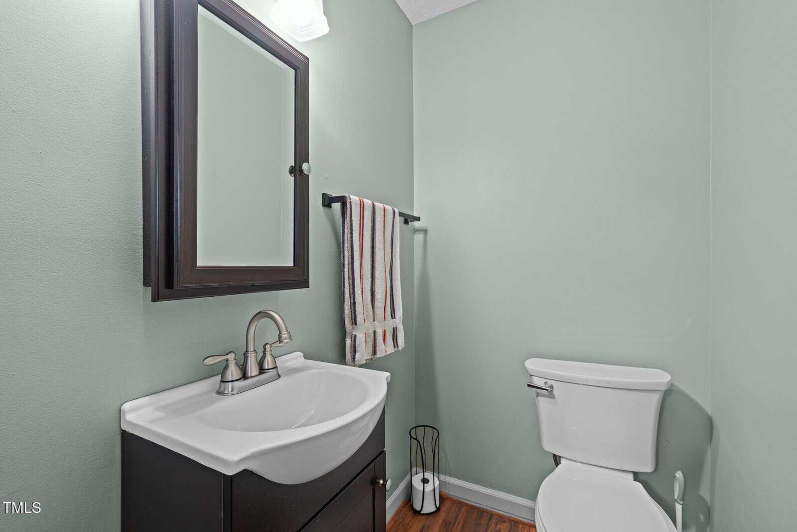 A bathroom with light green walls features a white sink with a dark brown cabinet, a silver faucet, and a mirror above. A striped towel hangs on a black towel rack. A white toilet is next to a black wastebasket on a wood floor.