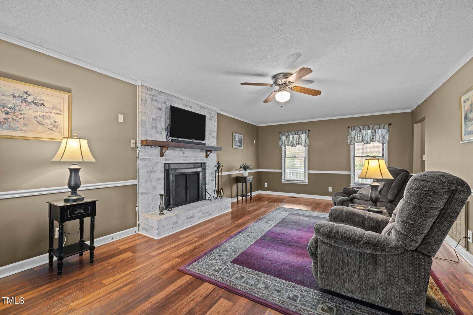 A living room with hardwood floors features two recliners and a light blue area rug. The room has a brick fireplace with a mounted TV above. Two lamps on side tables provide soft lighting, and two windows have floral curtains. A ceiling fan is overhead.