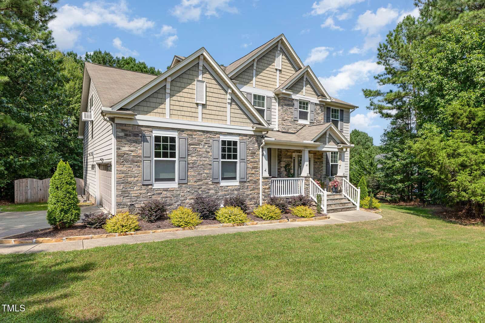 Two-story suburban house with stone and siding exterior, white trim, and a covered front porch. The house sits on a green lawn with small shrubs and trees, under a partly cloudy blue sky.