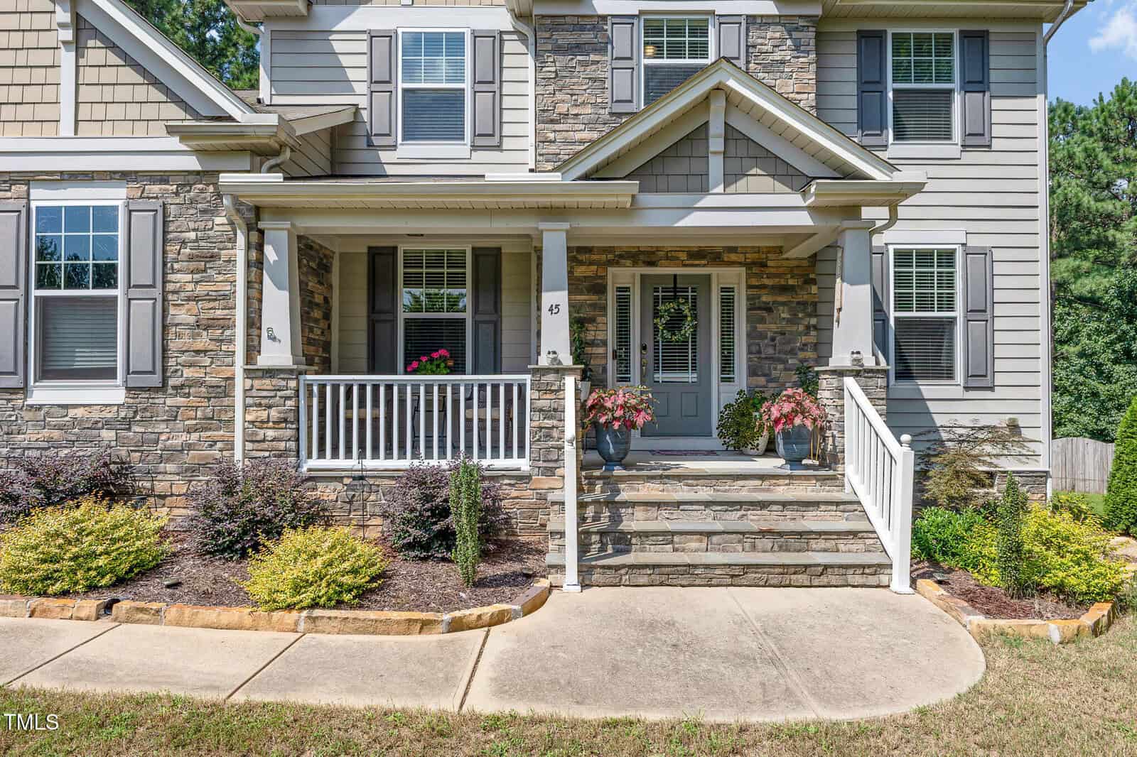 A two-story house with stone and gray siding exterior, white trim, front porch with white railings, potted plants, and steps leading to a double front door, surrounded by landscaped shrubs and grass.