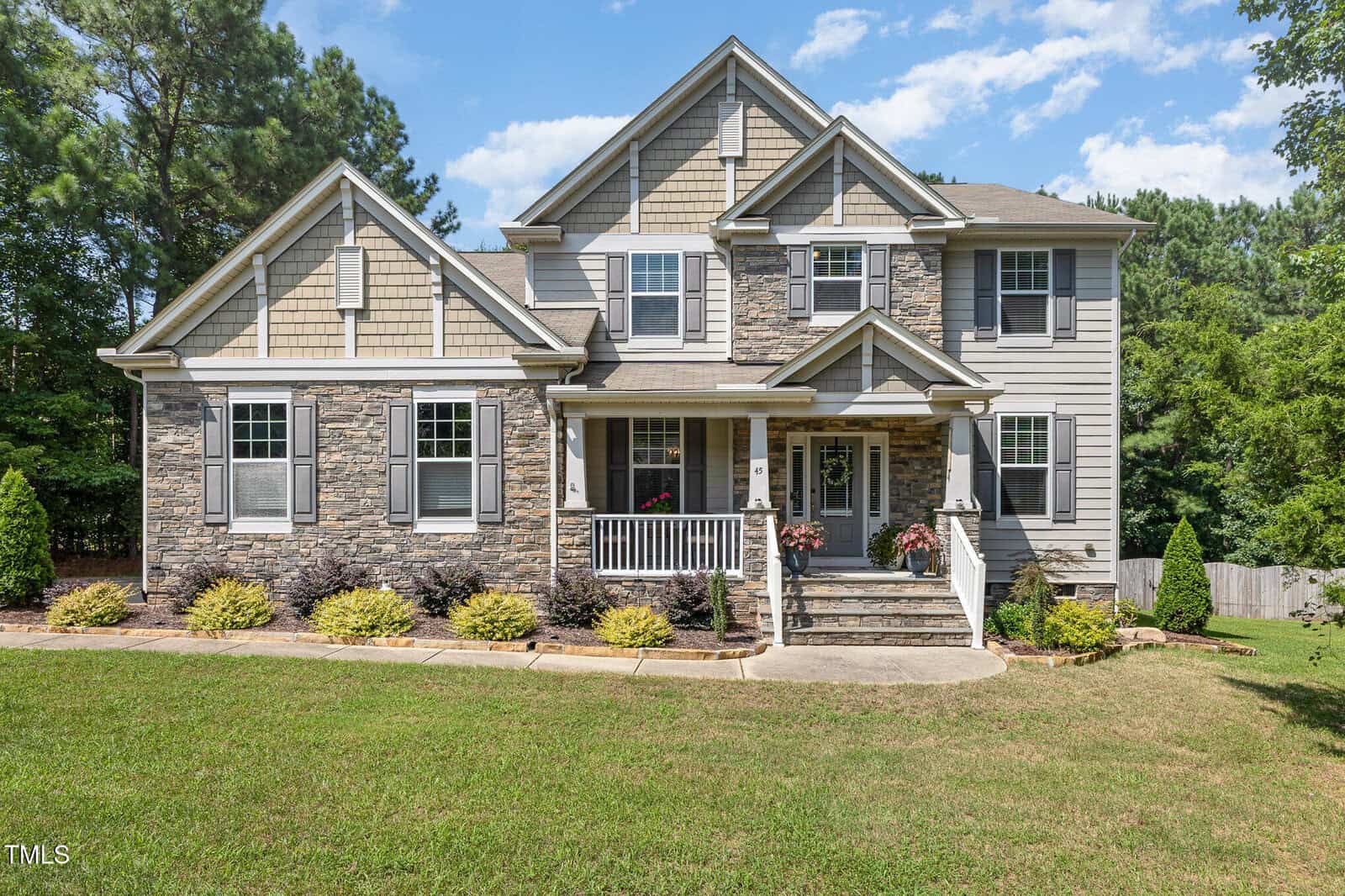 A two-story suburban house with stone and gray siding, white trim, a covered front porch, and landscaped yard with bushes and grass, surrounded by trees under a blue sky.