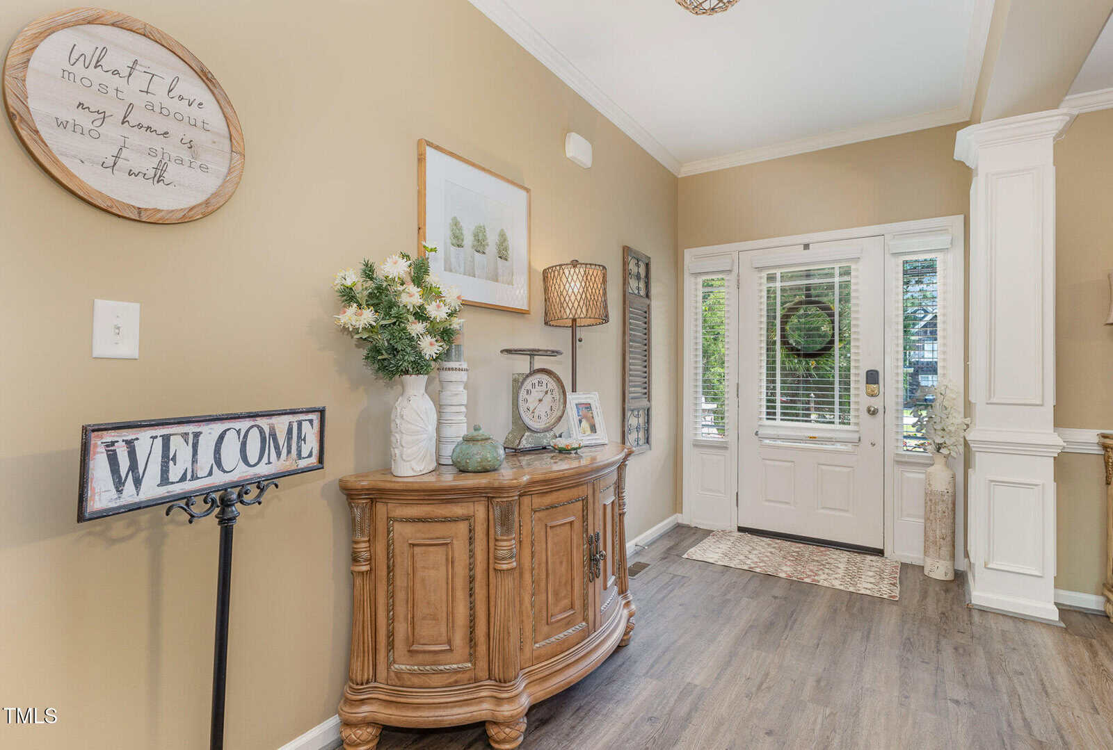 A warmly lit entryway with beige walls, a wooden console table decorated with flowers, framed art, a clock, and a "WELCOME" sign. White double doors with glass panels let in natural light.