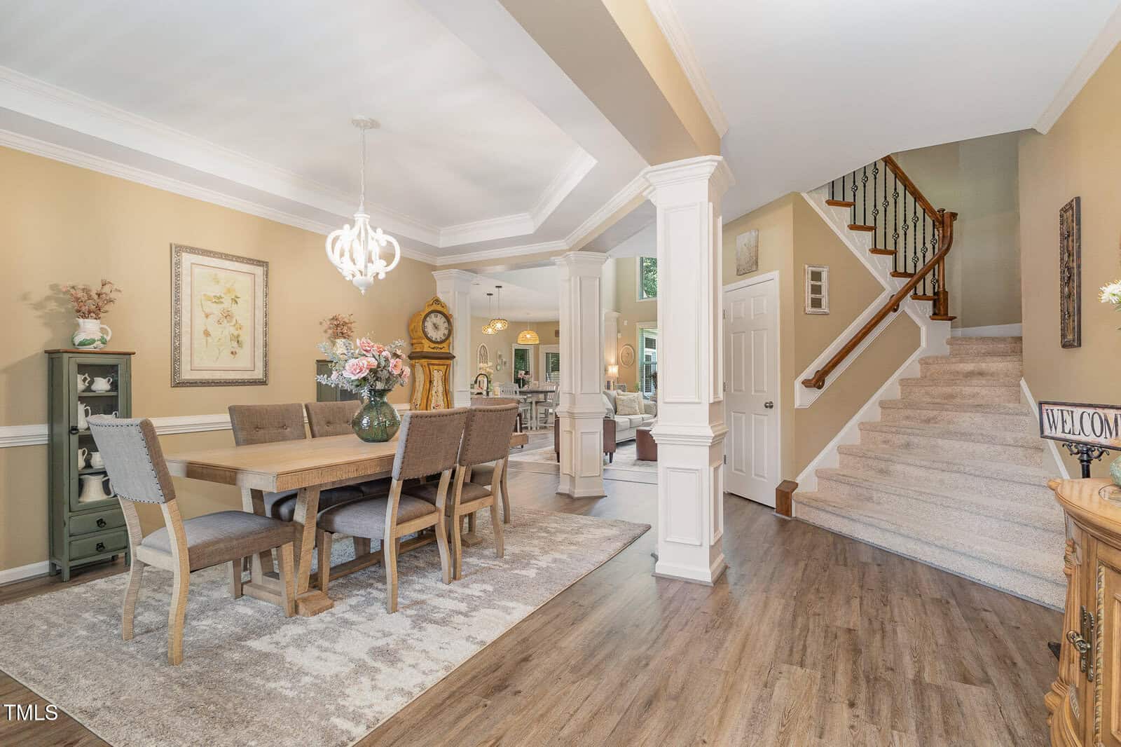 A dining room with a wooden table and six chairs on a rug, a light fixture above, and decorative items on cabinets. Stairs with a carpet runner lead upstairs. The space has hardwood floors and neutral-colored walls.