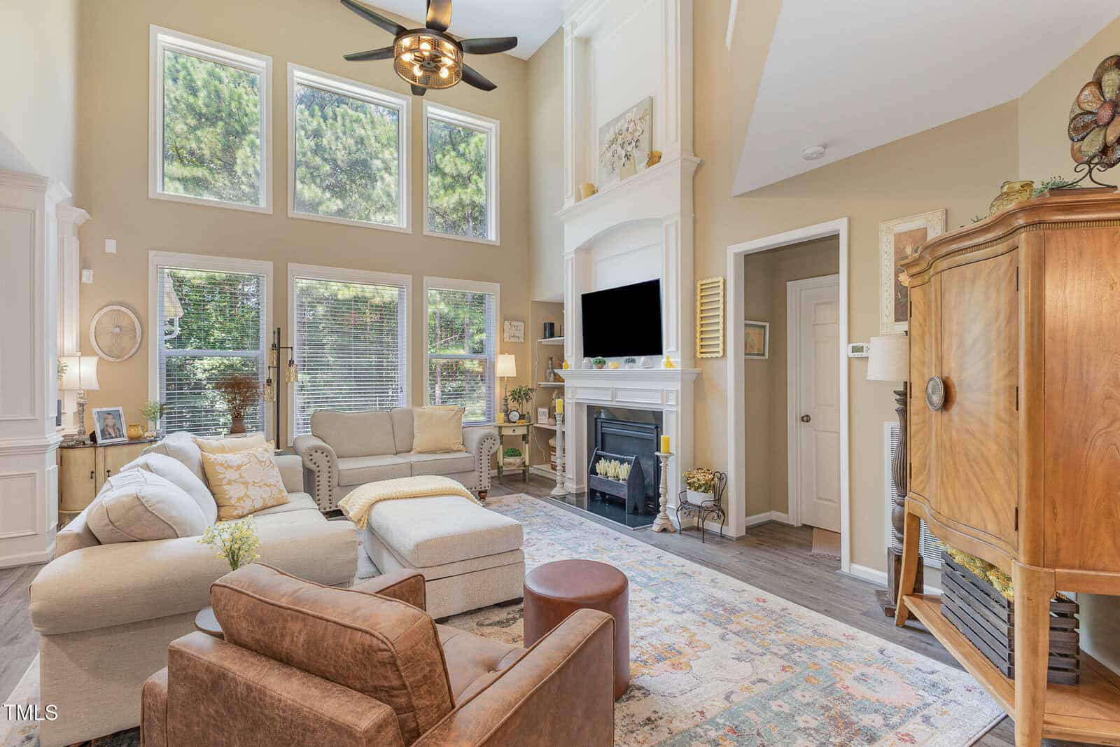 Bright living room with tall windows, neutral walls, a fireplace with a TV above it, light sofas, a brown armchair, wooden armoire, and a patterned rug. Sunlight fills the space, highlighting the cozy and tidy arrangement.