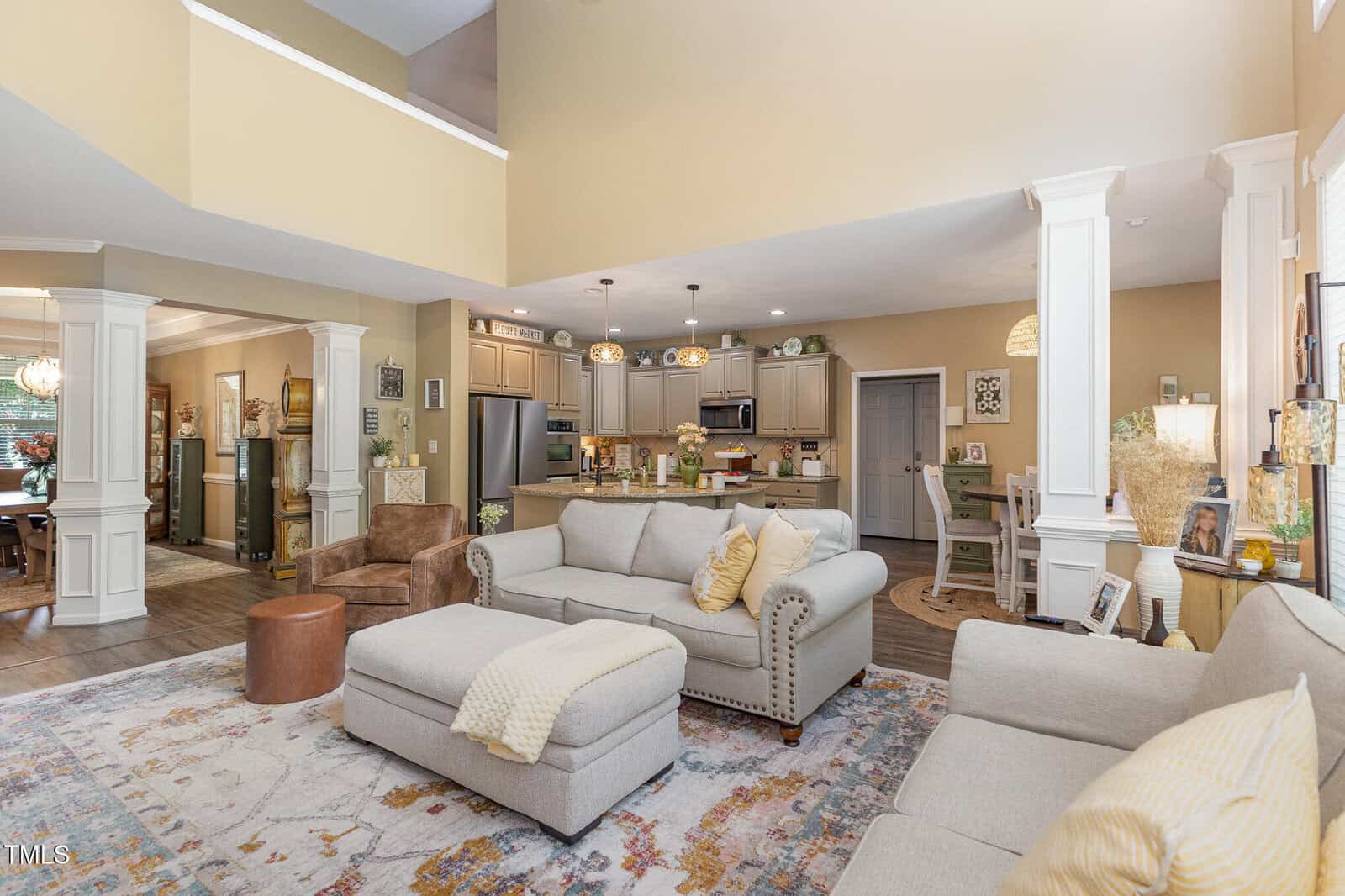 Open-concept living room and kitchen with beige walls, white trim, sofas, a patterned rug, kitchen island with barstools, pendant lighting, and various decor items. A dining area is visible in the background.