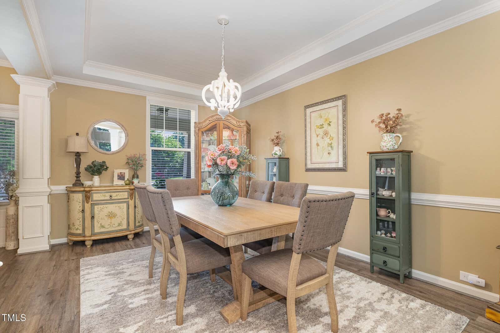 Dining room with a wooden table, six upholstered chairs, a chandelier, sideboard with decor, a glass cabinet, framed art, and beige walls. A large vase with flowers sits in the center of the table.