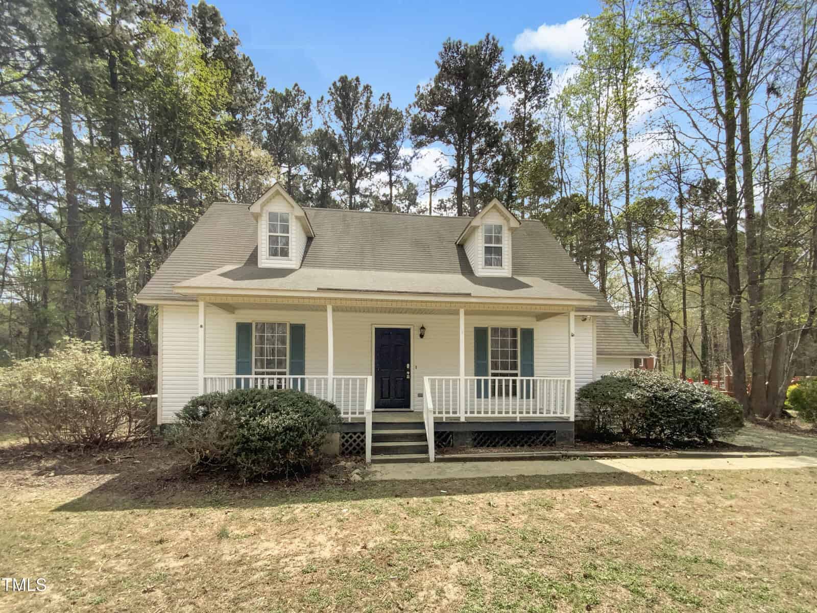 A small white house with a front porch, two dormer windows on the roof, green shutters, and bushes along the porch. The house is surrounded by trees and sits on a patchy lawn under a partly cloudy sky.