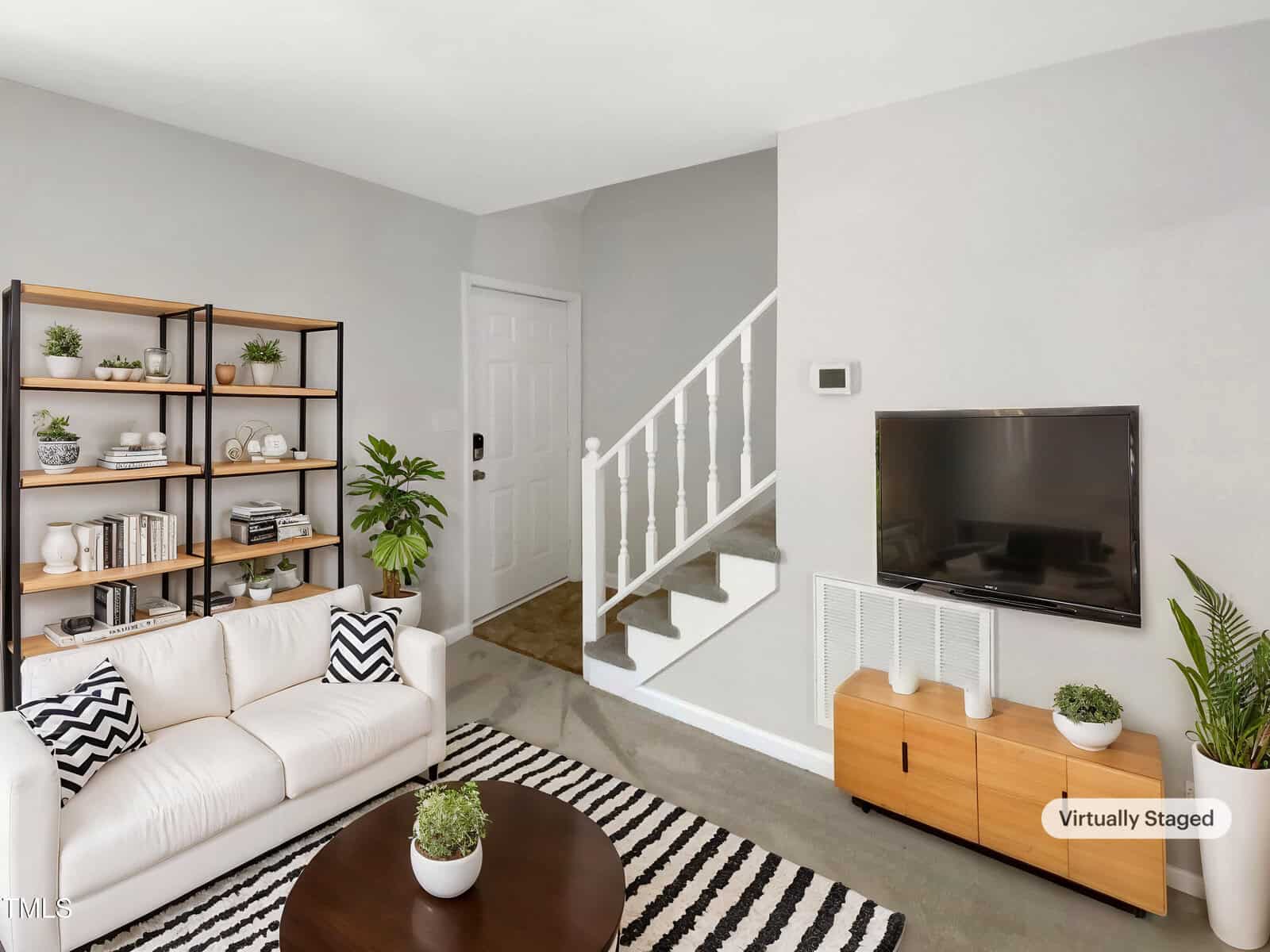 A modern living room with a white sofa, a wooden coffee table, a striped rug, a TV mounted on a light-colored wall, a wooden TV stand, a bookshelf with plants and decor, and a staircase in the background.