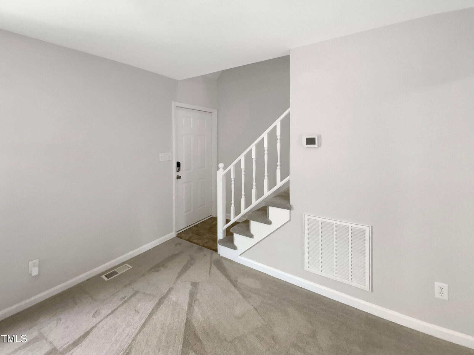 A small, carpeted room with light gray walls, a white front door, a white staircase with a handrail, a wall vent, thermostat, and electrical outlets. The area is clean and well-lit.