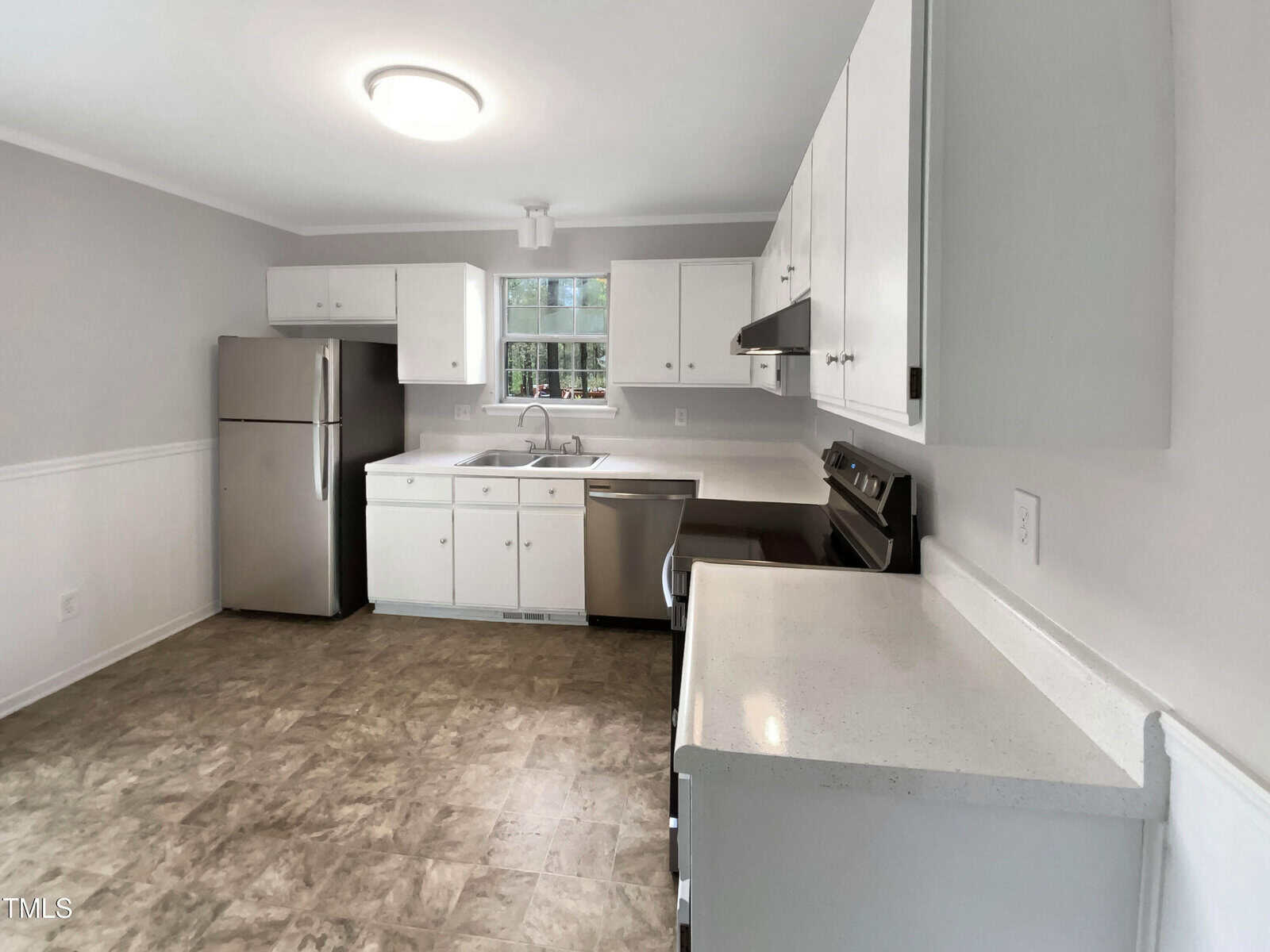 A kitchen with white cabinets, stainless steel refrigerator, dishwasher, and oven. There is a window above the sink, neutral-colored walls, and a light fixture on the ceiling. The floor has a brown patterned tile.