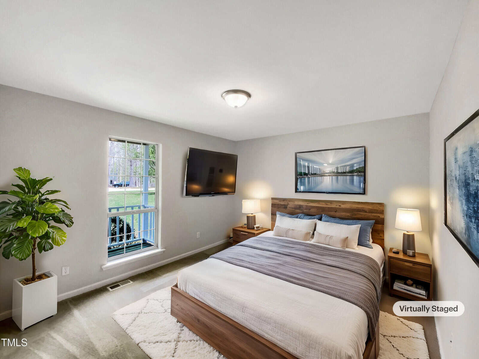 A bedroom with a large bed, two wooden nightstands, a wall-mounted TV, a potted plant by the window, and framed art above the bed. The room is well-lit with a window and a ceiling light. A "Virtually Staged" label is visible.
