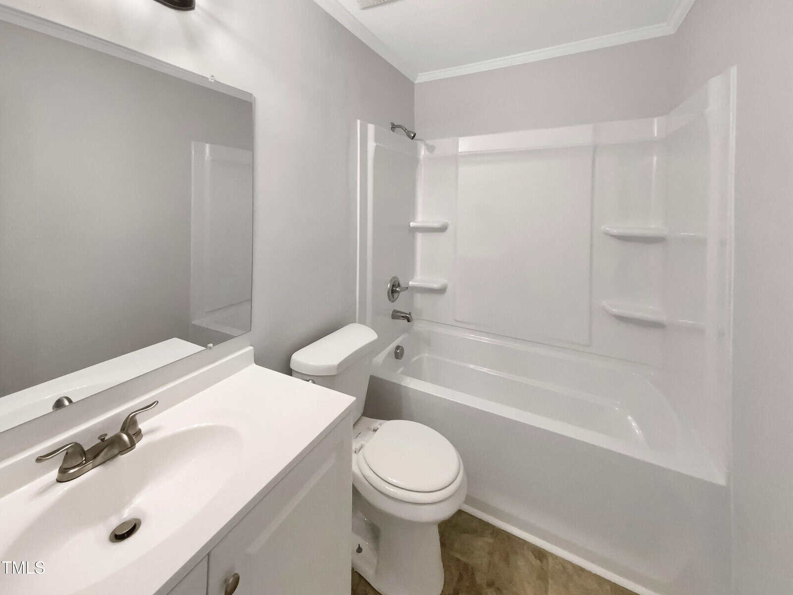 A small bathroom with a white sink and cabinet, a toilet, and a white bathtub-shower combination. There is a large mirror above the sink and a chrome faucet. The walls are light gray and the floor has a tan pattern.