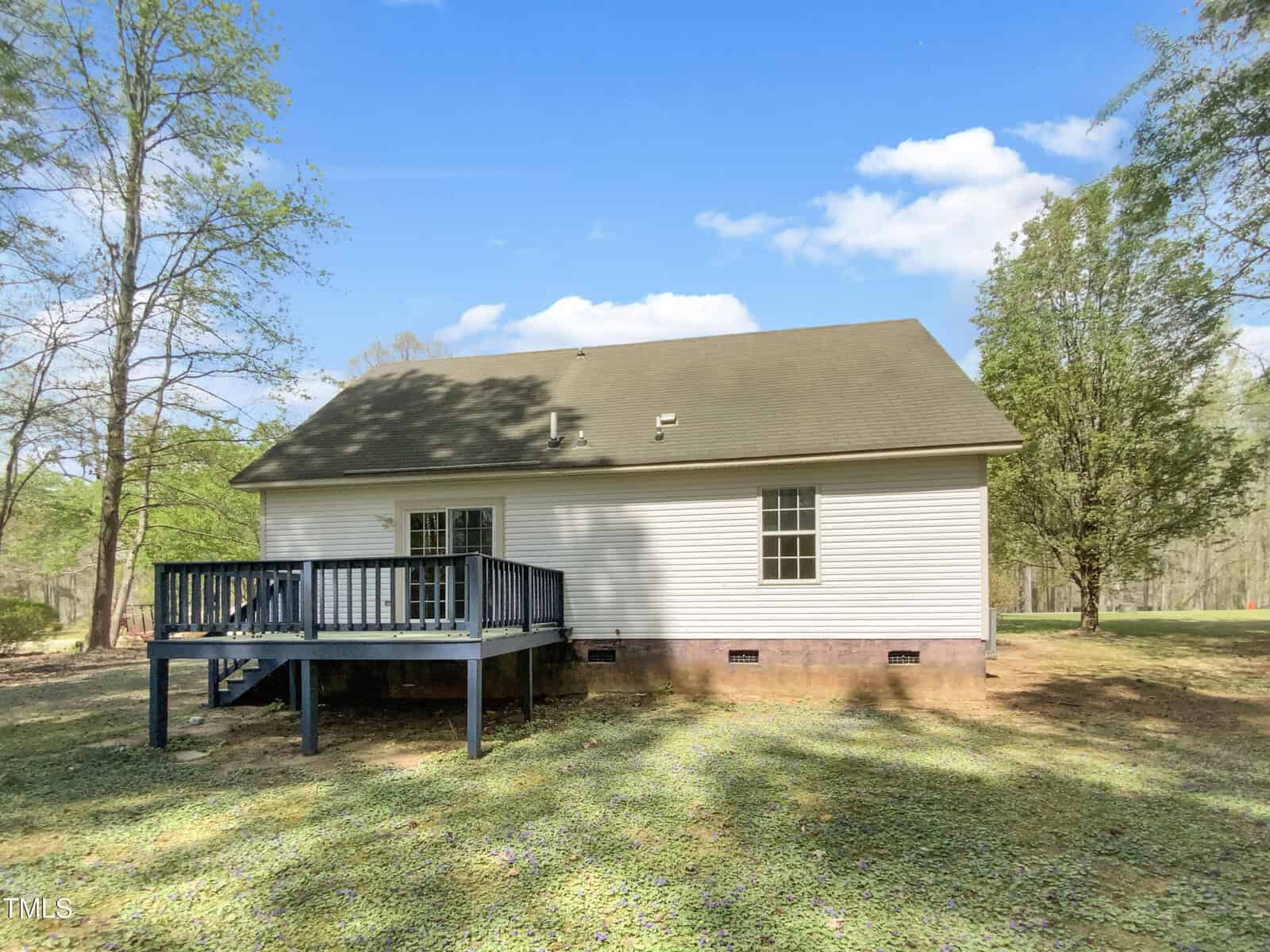 A white, single-story house with a gray shingled roof and a raised wooden deck in the backyard, surrounded by trees and grass under a clear blue sky.