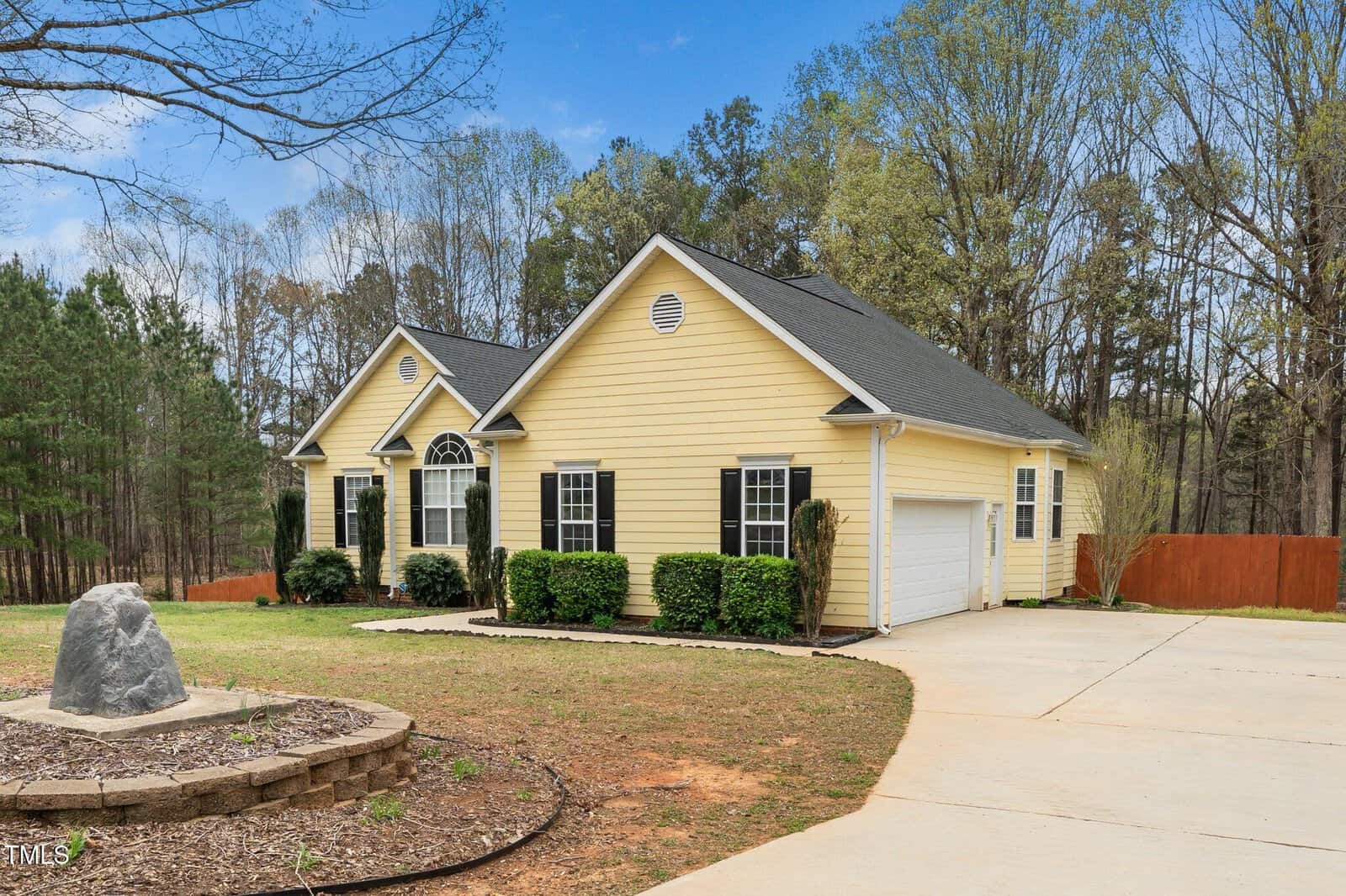 A single-story yellow house with a dark roof is set in a wooded area. There is a two-car garage and a concrete driveway. Bushes line the front of the house. A circular stone flowerbed with a large rock is in the foreground. Trees surround the property.