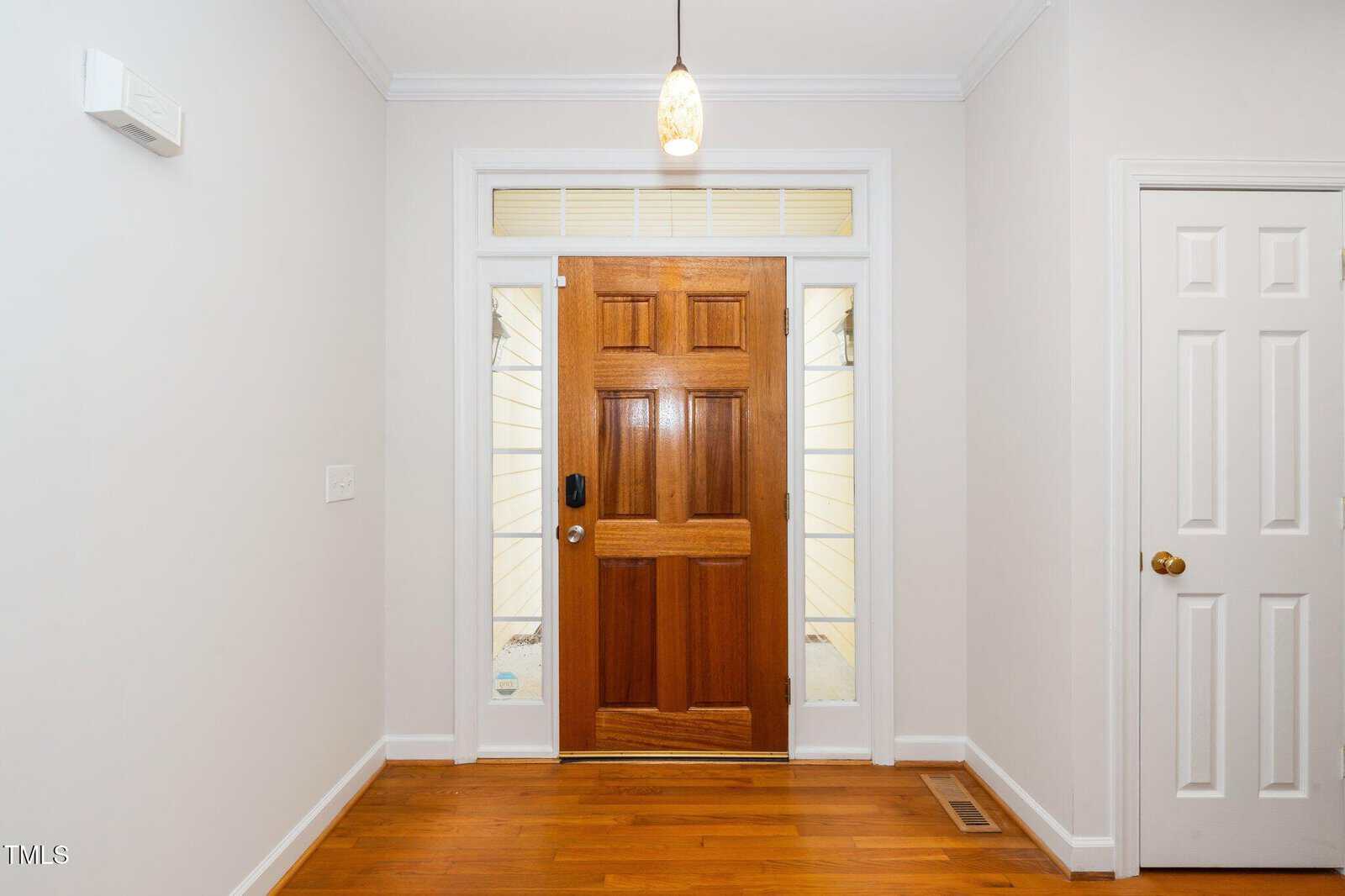A hallway with light-colored walls features a wooden front door with a rectangular window above it. The space includes a pendant light on the ceiling, a white closed door on the right, and a glossy wooden floor.