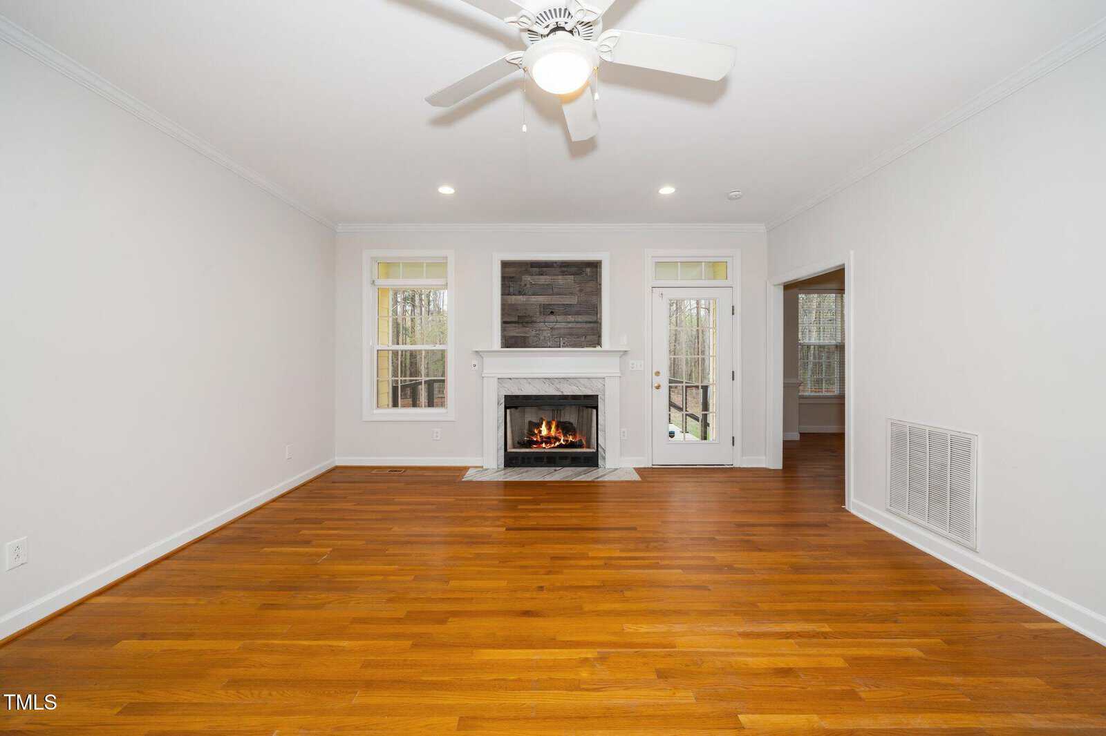 A spacious living room with hardwood floors and white walls. A lit fireplace with a dark wood mantel is centered on the back wall. Large windows flank the fireplace. A white ceiling fan is overhead, and a doorway leads to another room on the right.