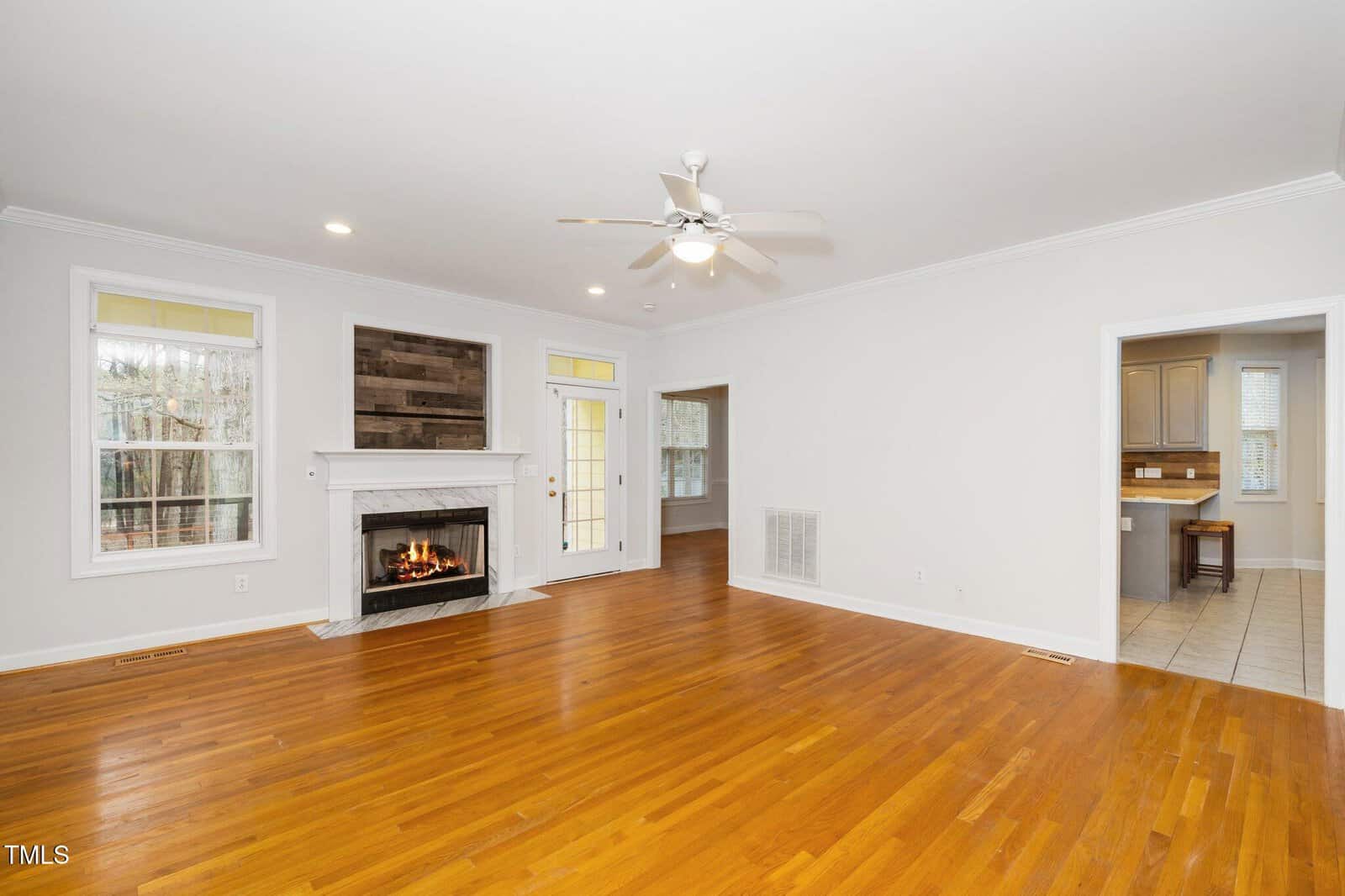 Spacious living room with hardwood flooring, a white fireplace with a wooden mantel, and a ceiling fan. Large windows and a glass door provide natural light. The room opens to a kitchen area with tile flooring on the right.