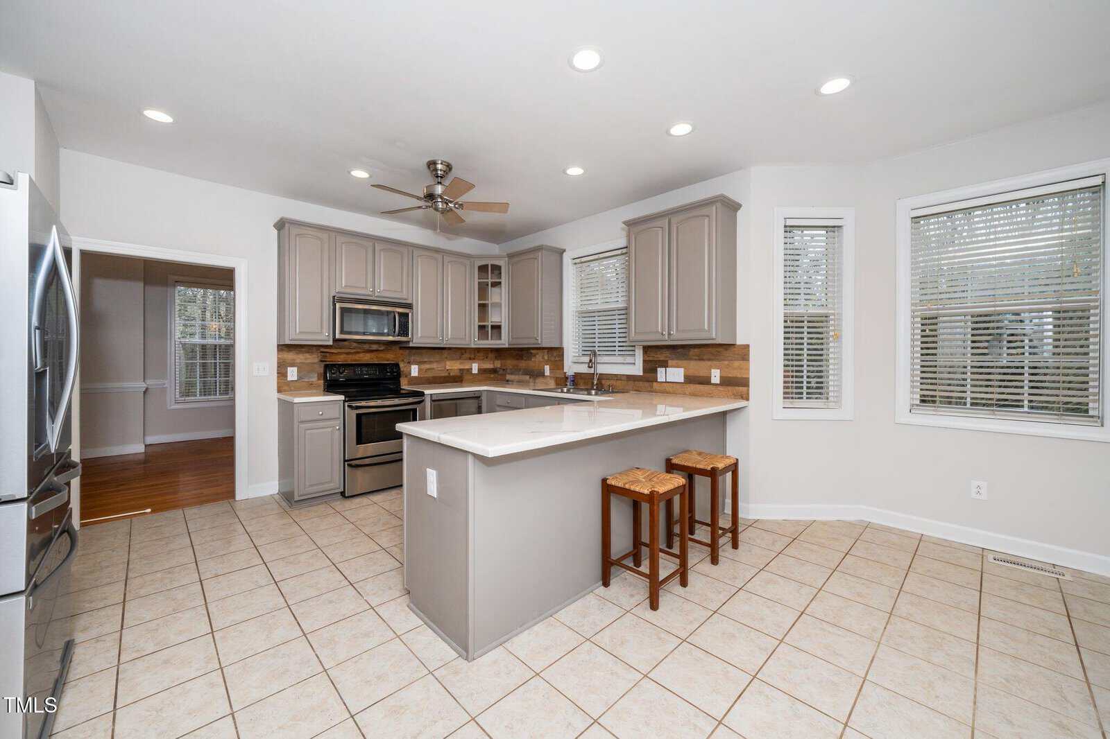 Modern kitchen with beige cabinets, stainless steel appliances, and a central island with a white countertop and two wooden stools. The floor is tiled, and there are three windows with blinds. Ceiling fan and recessed lighting are visible.