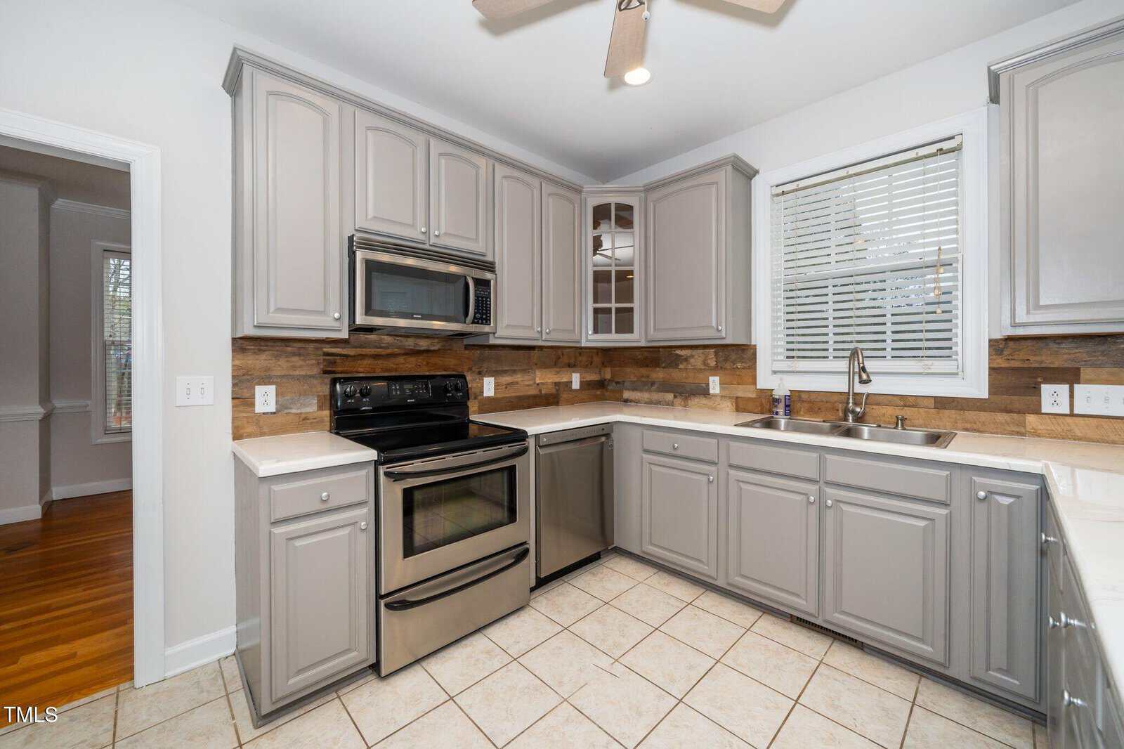 A kitchen with gray cabinets, stainless steel appliances, and a double-basin sink. The countertop is light-colored, and the backsplash has a wood-like texture. A window with blinds is above the sink, and the floor is tiled.