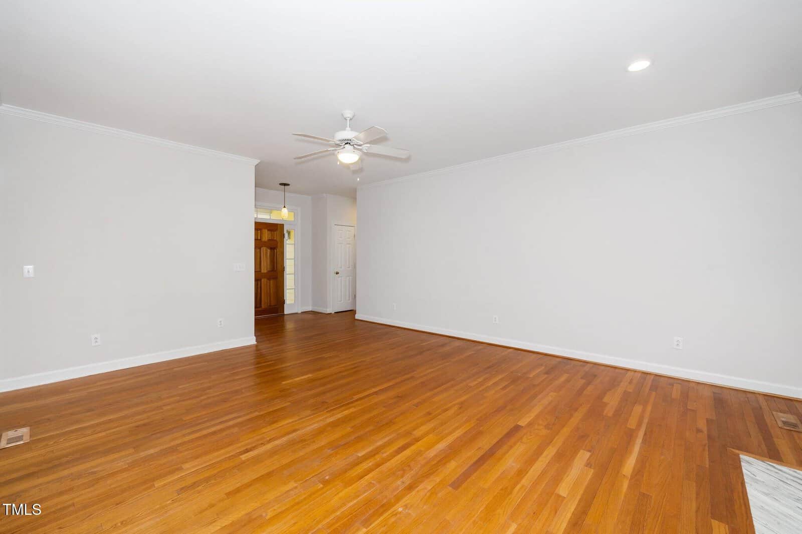 An empty room features polished hardwood floors, white walls, and a white ceiling with a ceiling fan and recessed lighting. A doorway leads to a hallway with a visible wooden door. No furniture is present.