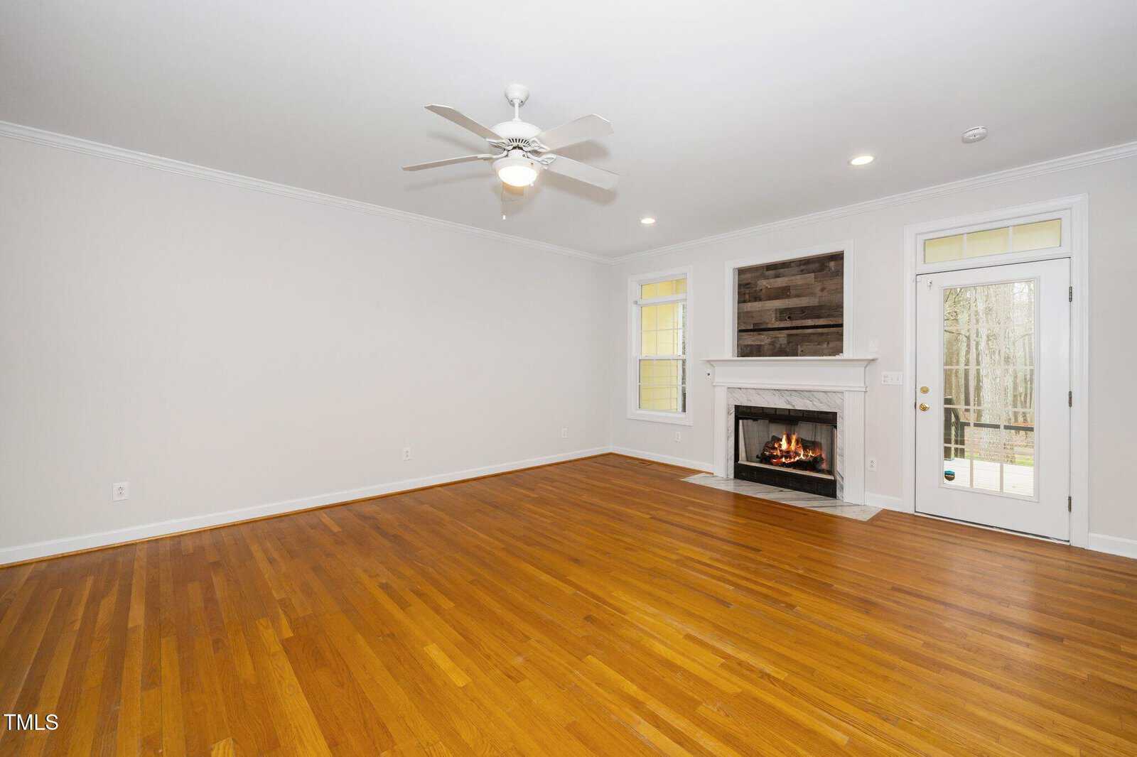 A spacious, empty living room with polished wooden floors. The room features a ceiling fan, white walls, recessed lighting, and a fireplace with a wooden mantel. Large windows and a glass door provide natural light.