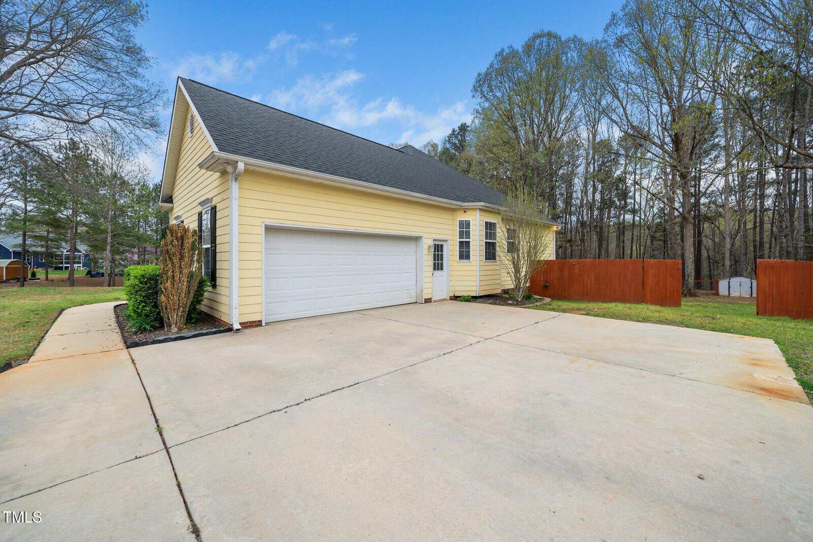 Single-story house with yellow siding and a dark gray roof, features a two-car garage. The driveway is large and made of concrete. A wooden fence borders the backyard, and trees surround the property, with a clear blue sky overhead.