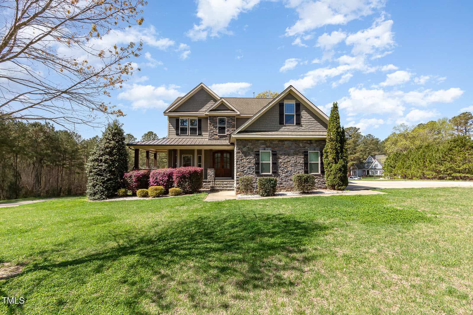 A two-story stone and wood house with a wrap-around porch sits on the spacious green lawn of 3034 East Bay Court in Creedmoor. It is surrounded by shrubs, trees, and a clear blue sky with scattered clouds. A driveway is visible to the right.