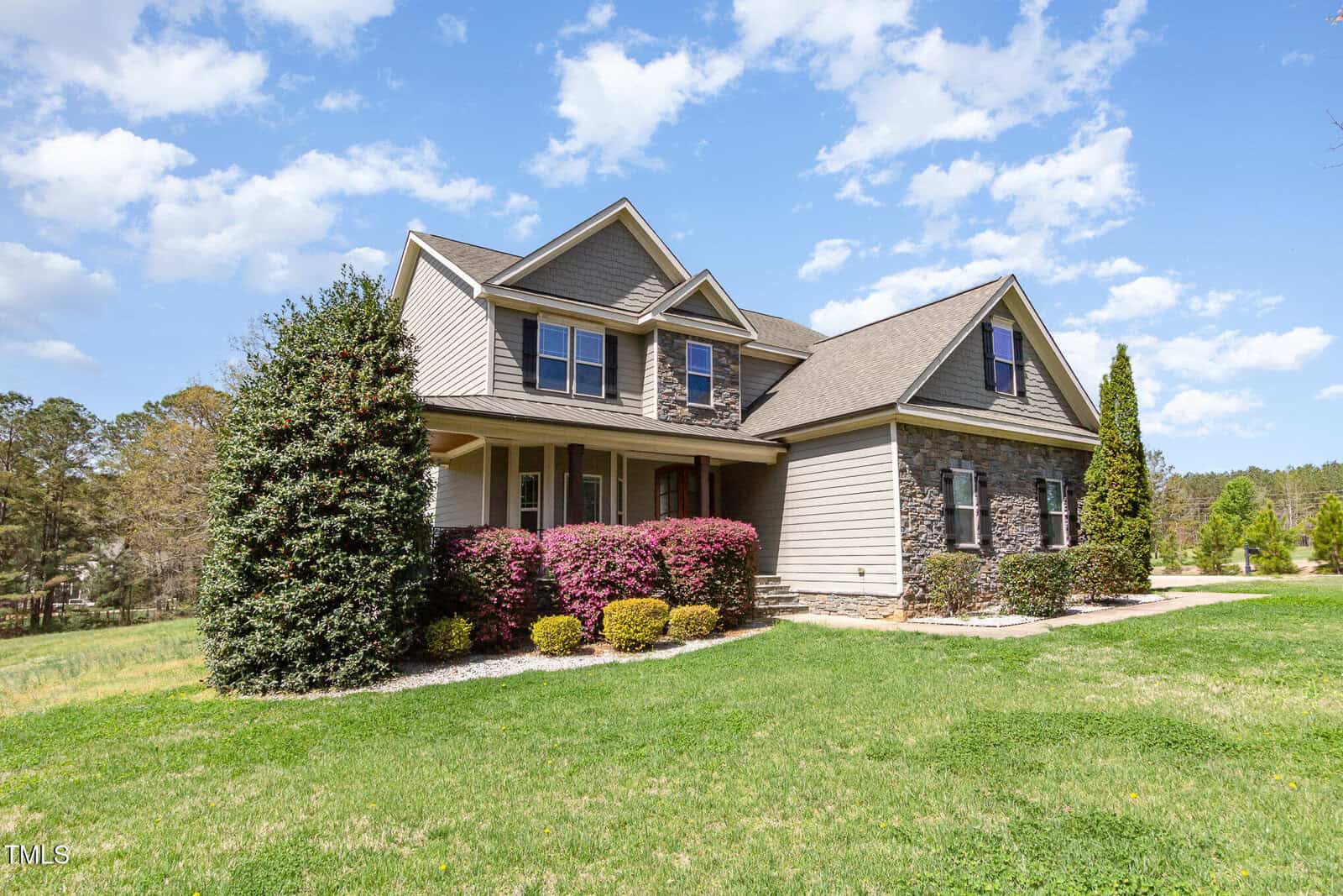 A two-story house with a mix of gray siding and stone exterior stands amidst a landscaped yard. There are trimmed bushes and a well-maintained lawn in the foreground, with a backdrop of trees and a partly cloudy sky.