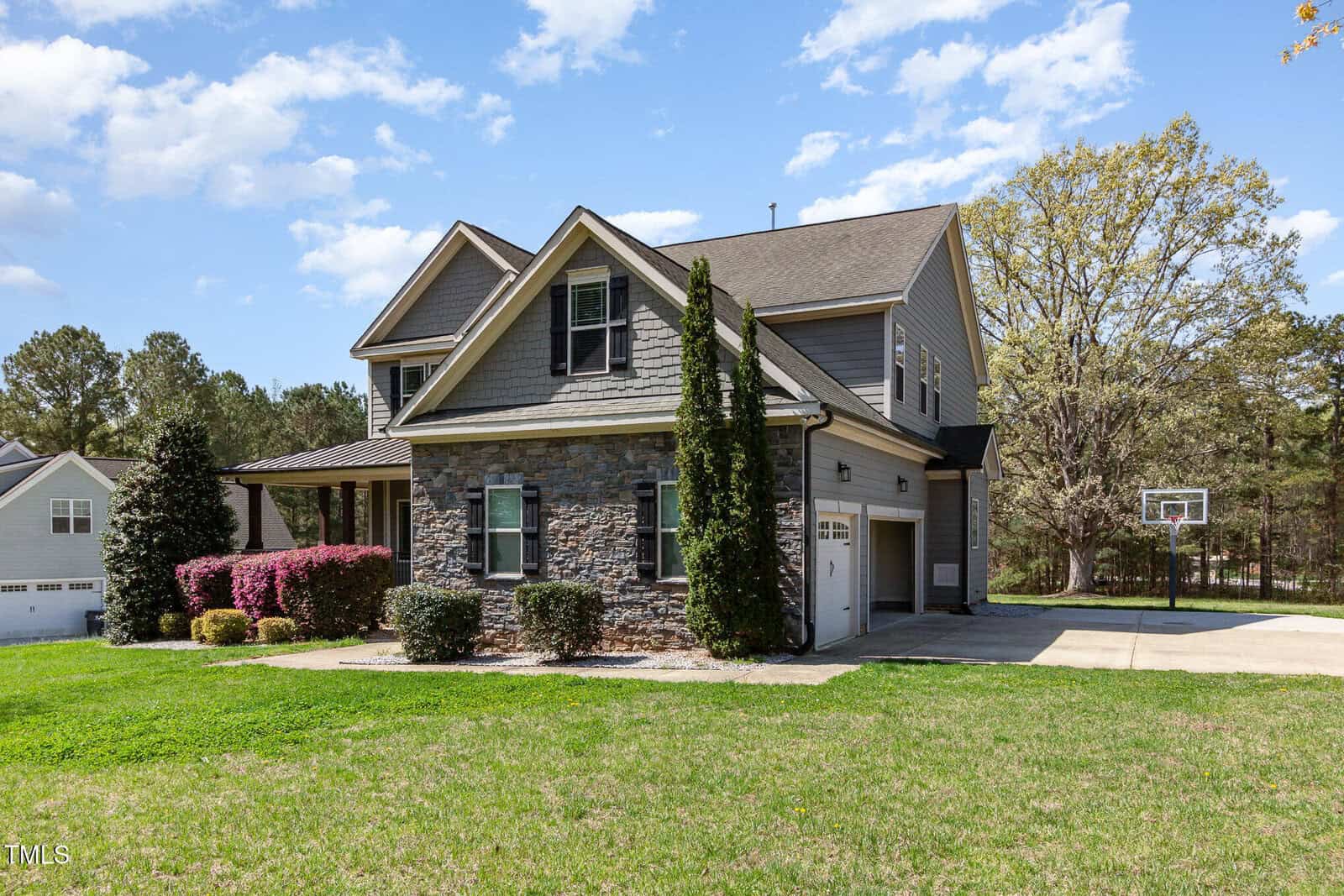 A two-story house with gray siding and stone accents in front, featuring a two-car garage. There are shrubs and a tree in the front yard, with a basketball hoop visible in the driveway on the right. The sky is partly cloudy.
