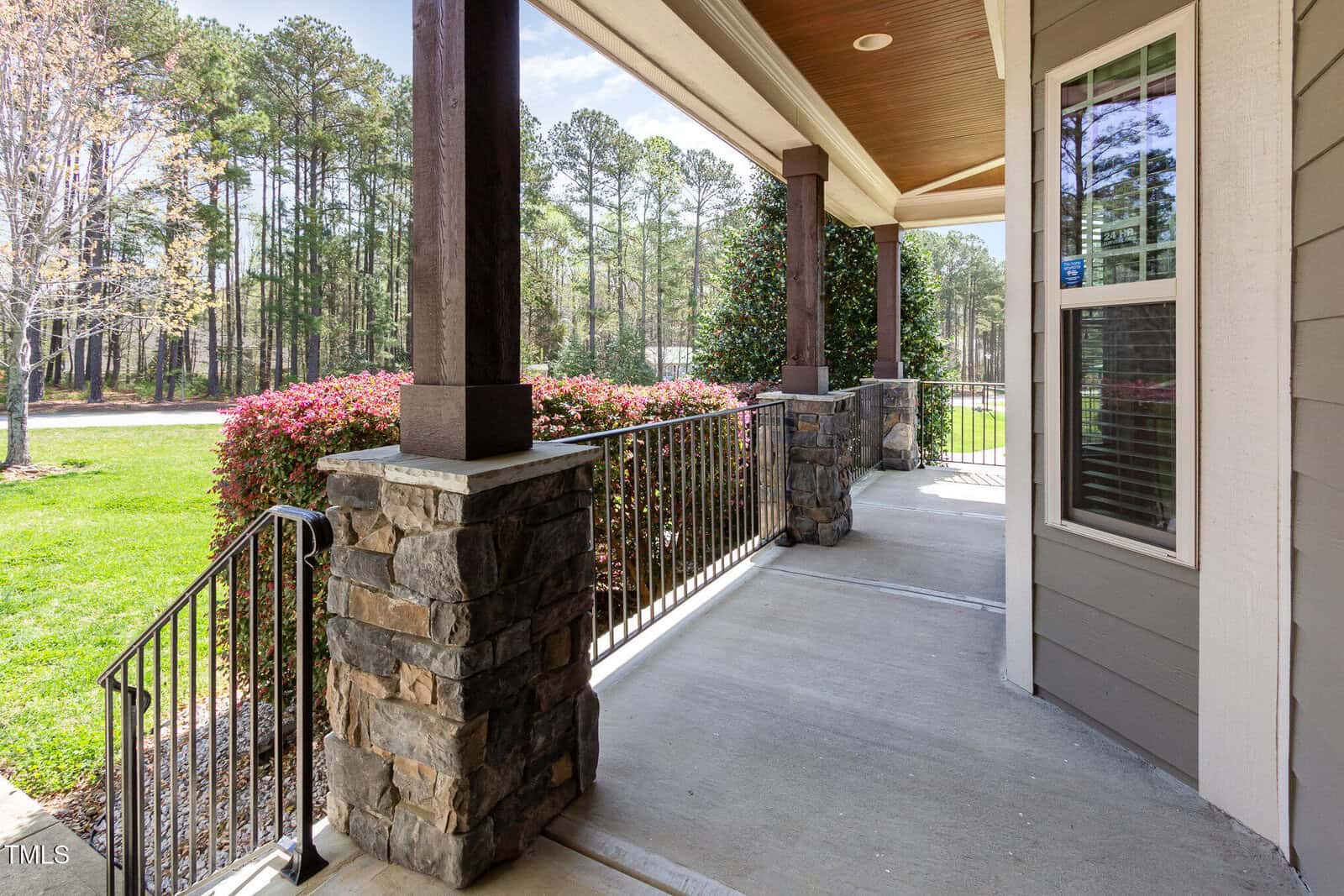 A covered porch with stone columns and metal railings overlooks a grassy yard and a row of trees. There are blooming pink shrubs next to the porch. The house exterior features beige siding and windows with white trim.