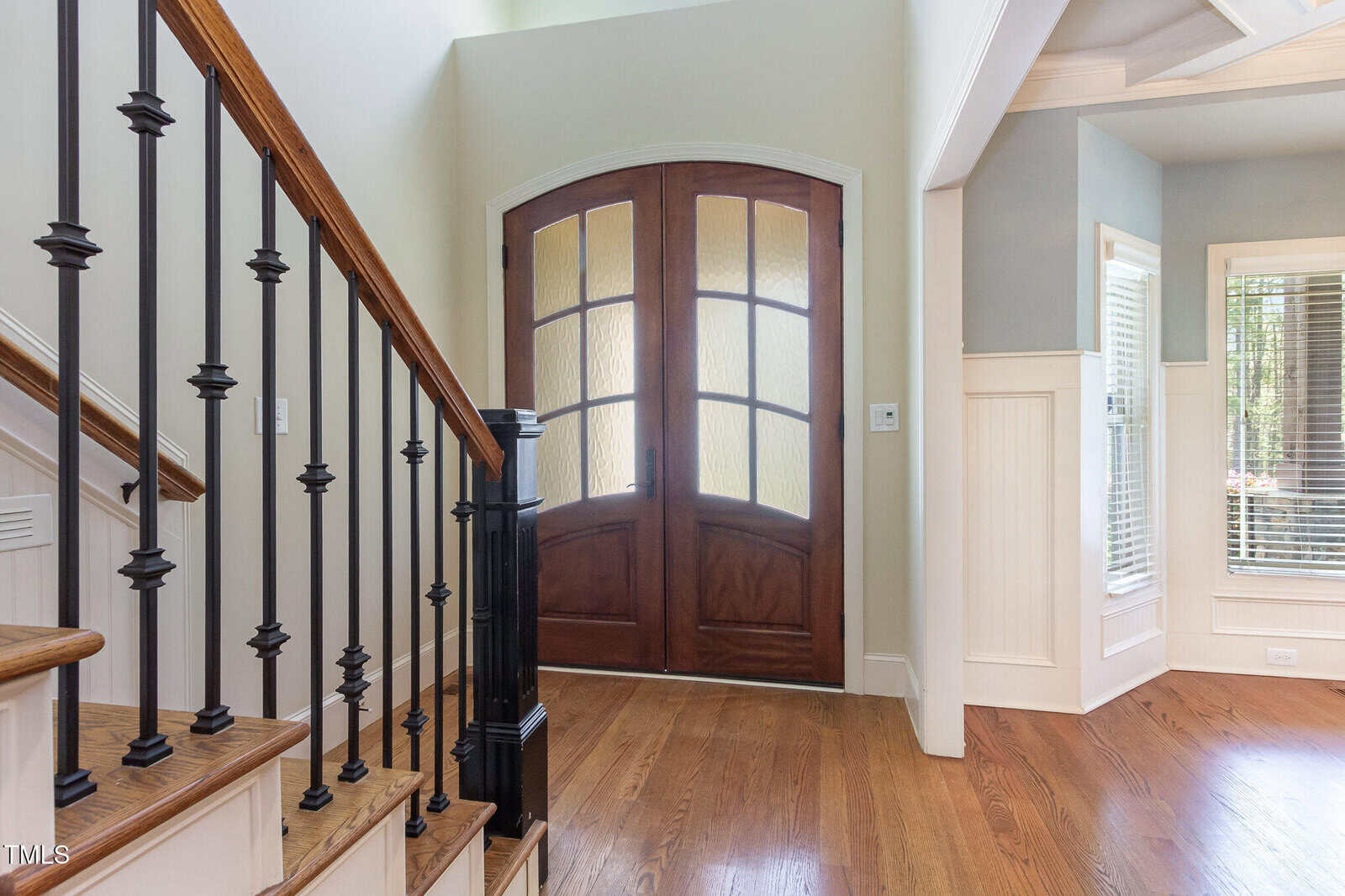 A foyer with a wooden staircase on the left, featuring black metal balusters. In the background, there are double wooden doors with glass panels. The floor is hardwood, and there is wainscoting on the right wall, leading to another room with large windows.