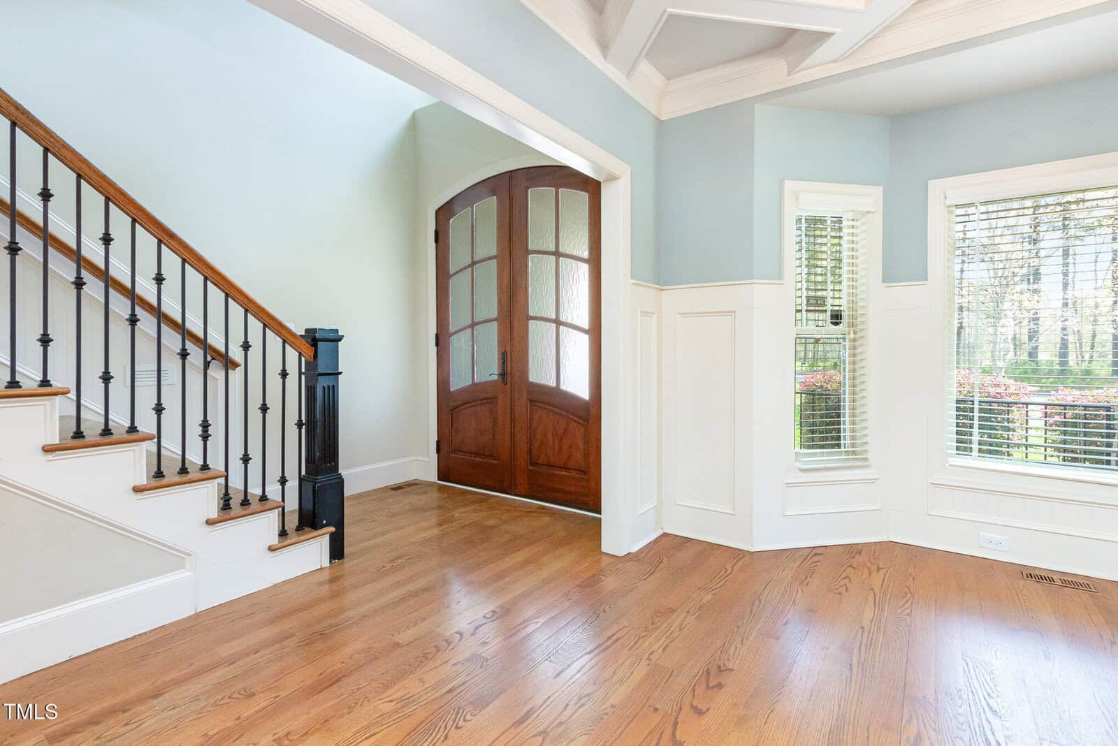 A spacious entryway featuring a wooden front door with glass panels, light blue walls, and white wainscoting. A wooden staircase with black metal railings is on the left, and a window with white blinds offers a view of greenery outside.
