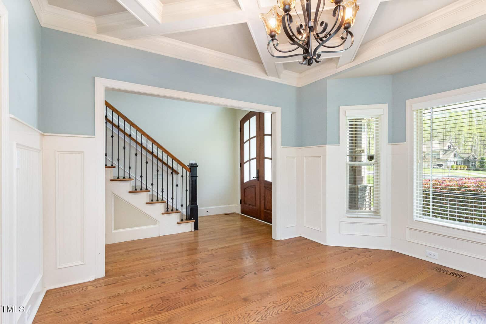 A spacious, empty room with light blue and white walls, featuring a wooden floor. There's a coffered ceiling with a chandelier, large windows with blinds, and an open doorway leading to a staircase with wooden steps and a railing.