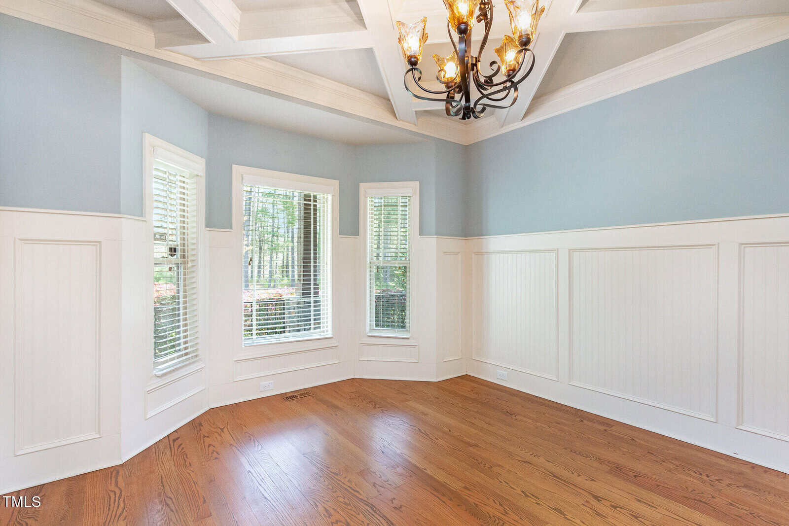 A bright, empty room with light blue walls and white wainscoting. It features a wooden floor, three tall windows with blinds, and a chandelier hanging from a coffered ceiling. The windows offer a view of trees outside.
