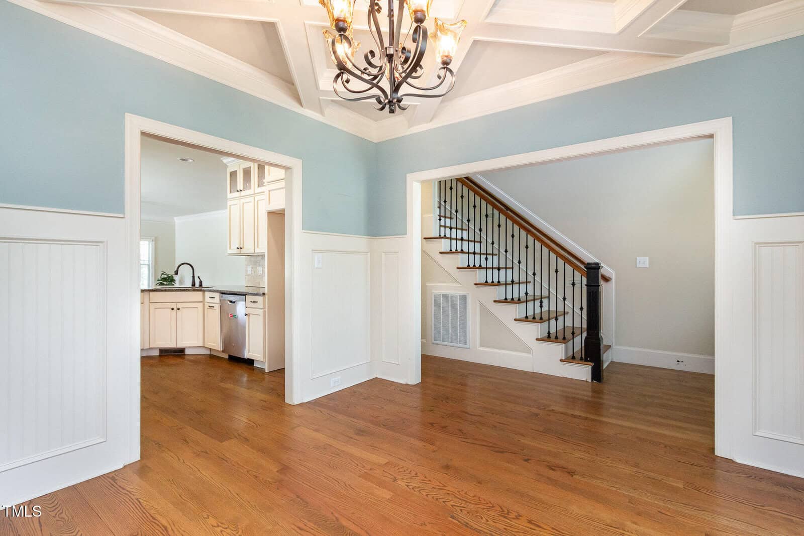 A spacious room features wooden floors and light blue walls with white wainscoting. Two open doorways lead to a kitchen and a staircase. A chandelier hangs from the coffered ceiling. A staircase with wooden railings is visible in the background.