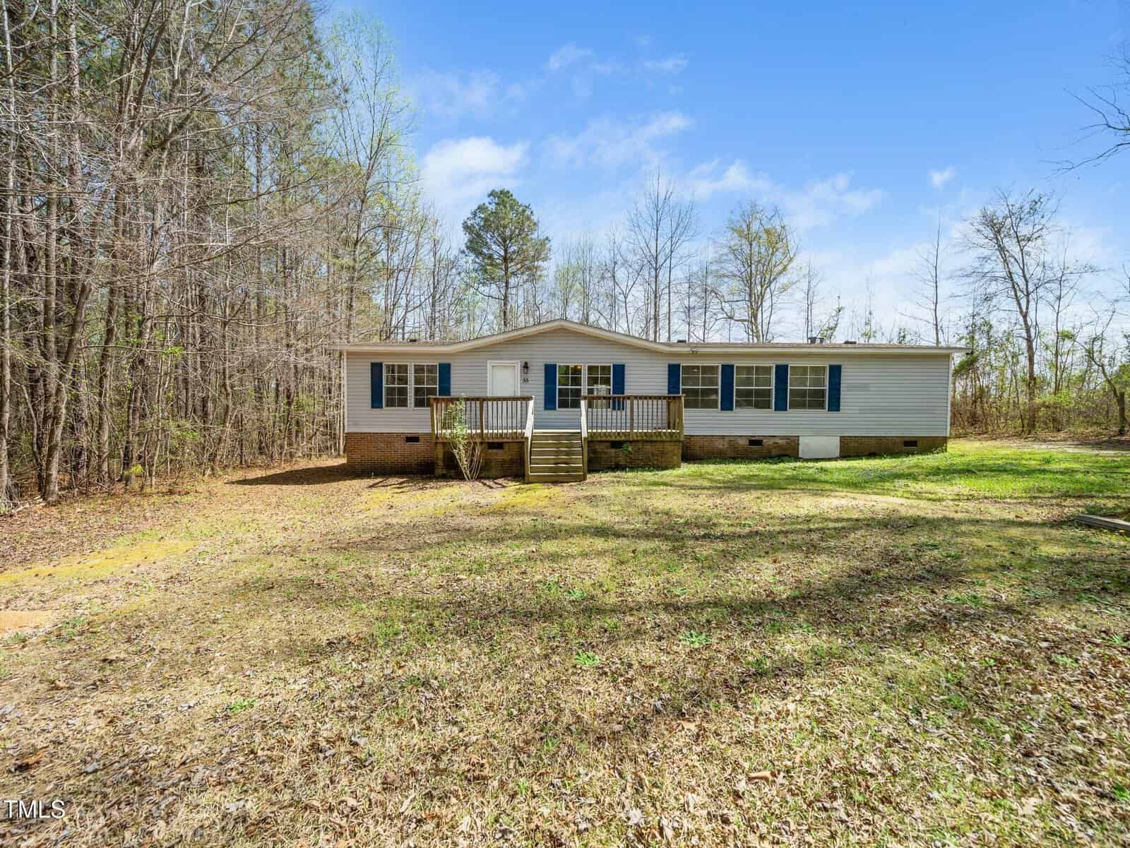 A single-story, light-colored house with blue shutters and a front porch, set on a grassy lot surrounded by tall trees under a partly cloudy sky.