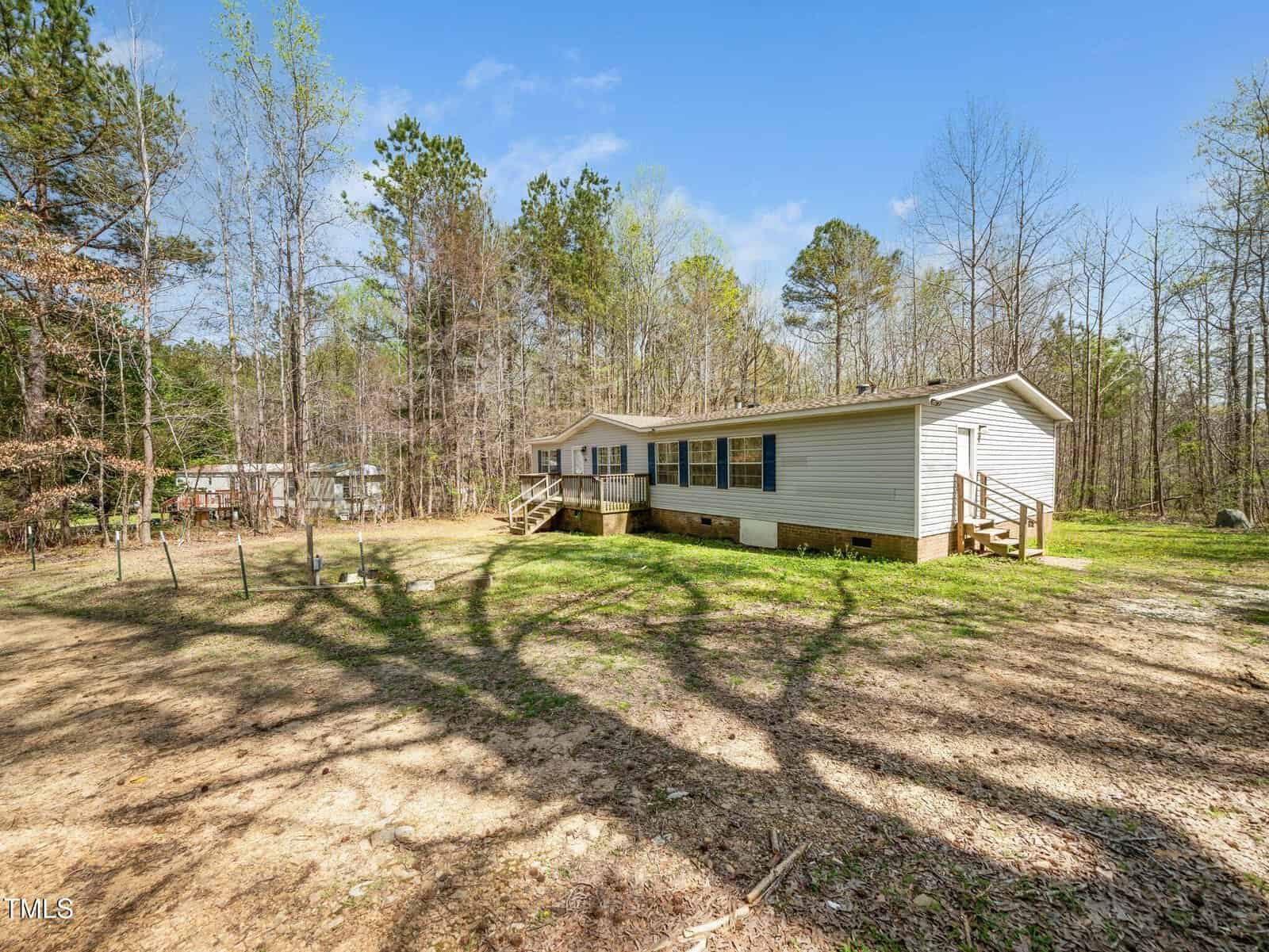A single-story manufactured home with white siding sits on a grassy, lightly wooded lot. There are stairs leading up to the front and side entrances. Tall trees surround the property, casting long shadows on the ground.
