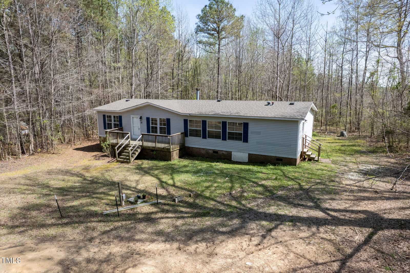 A single-story manufactured home with beige siding and a shingled roof is surrounded by trees. The house has front steps leading to a small porch and sits on a grassy, partially shaded lot.
