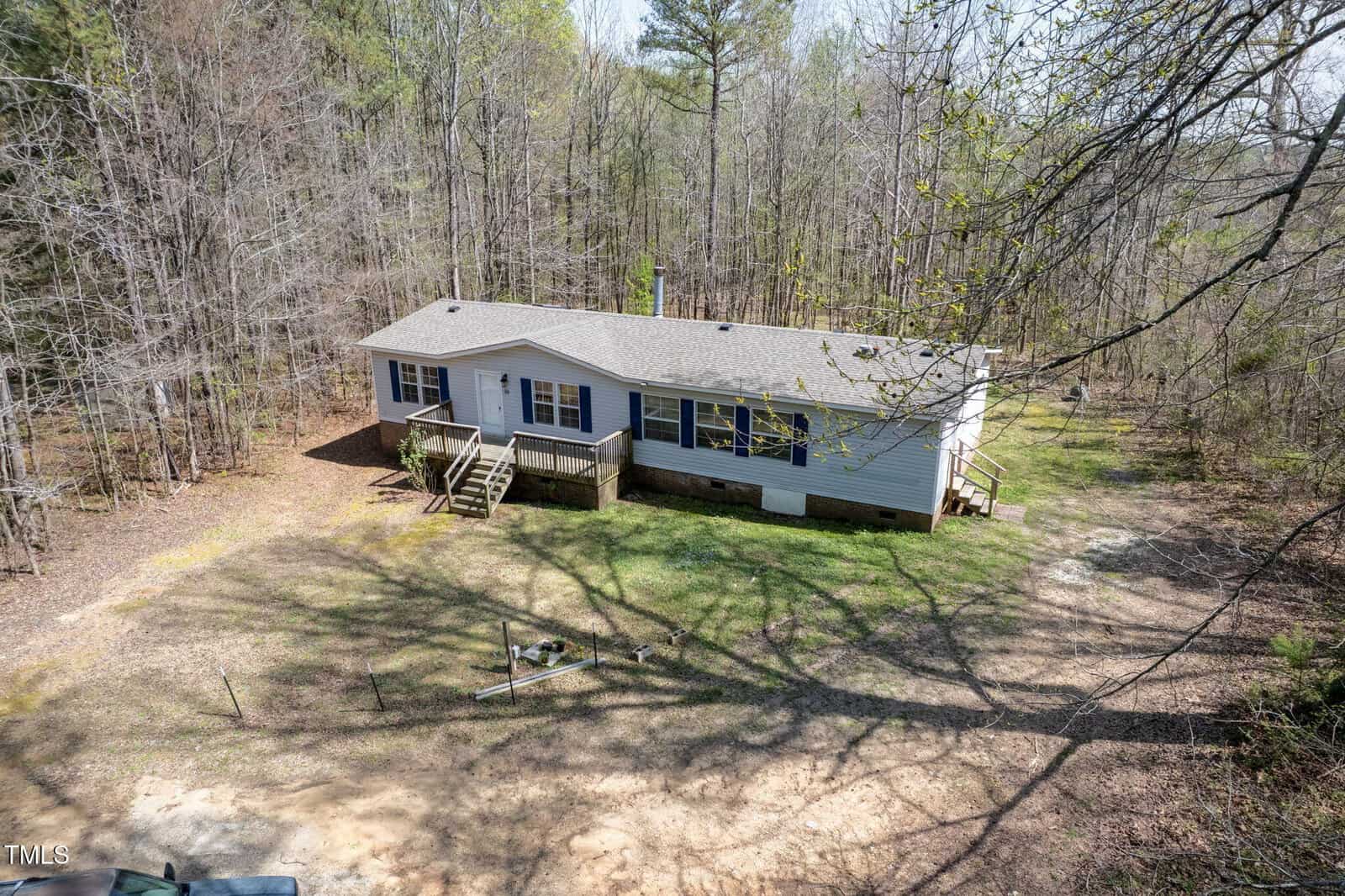 A single-story, light-colored house with a front porch and stairs is set in a clearing surrounded by trees. There is a small fenced area in the front yard, and the ground is partially covered with grass and dirt.