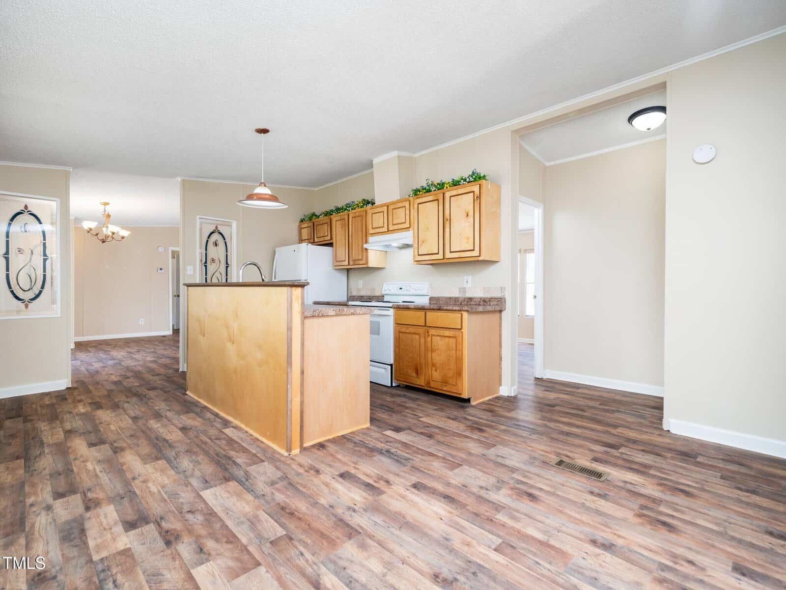 Open kitchen and dining area with wood floors, light wood cabinets, white appliances, beige walls, and decorative greenery above cabinets. A chandelier hangs in the dining space. The room is brightly lit with natural light.